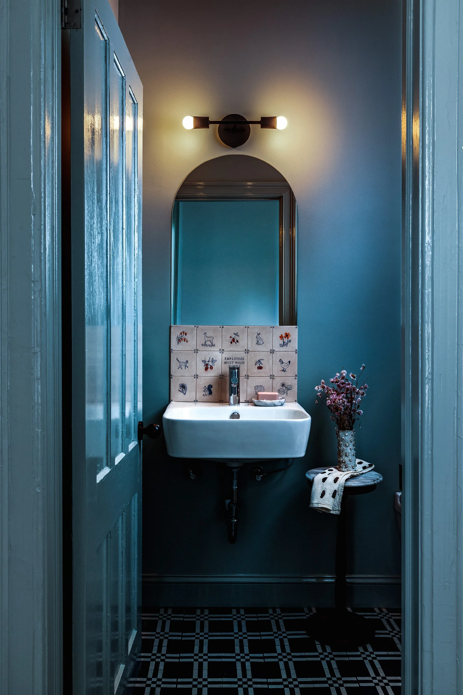 A small powder room featuring deep blue painted walls, a wall-mounted sink with illustrated backsplash tiles, an arched mirror, and a checkered floor.