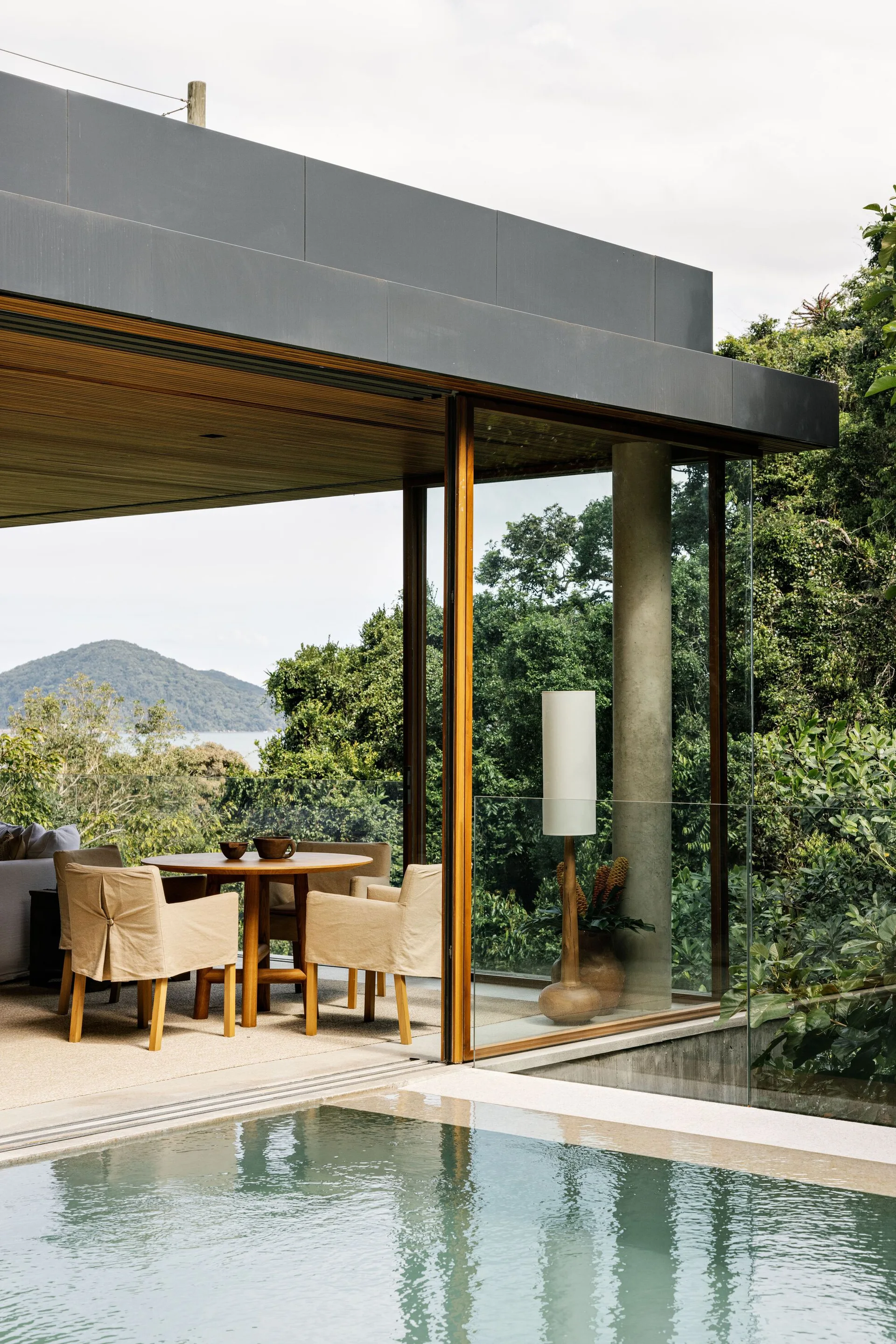 A poolside patio features a round wooden dining table with slipcovered chairs beneath a wood slat ceiling next to a swimming pool.