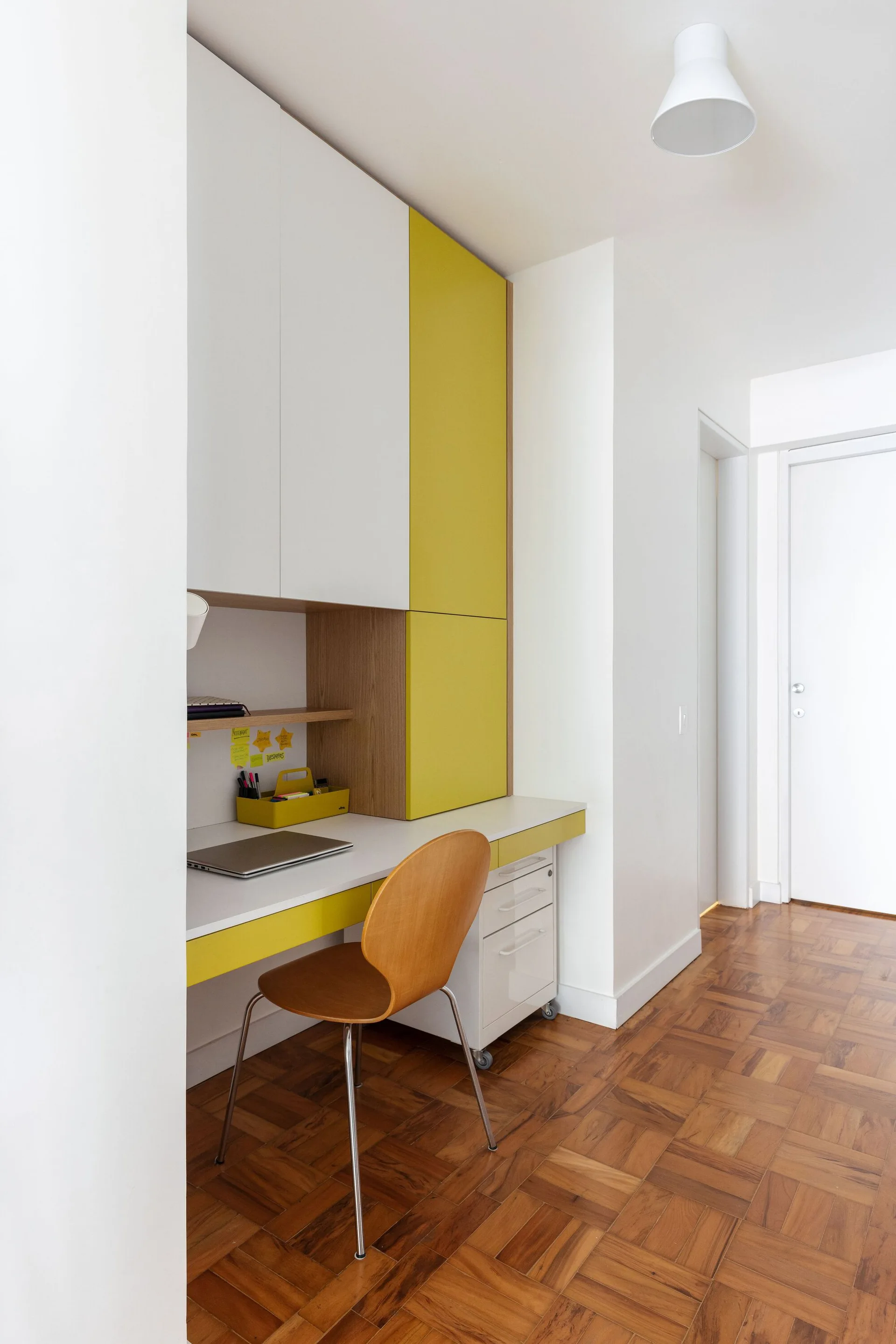 A modern built-in desk features white and yellow cabinets, a wooden desk chair, a white rolling drawer unit, and parquet wood floors.
