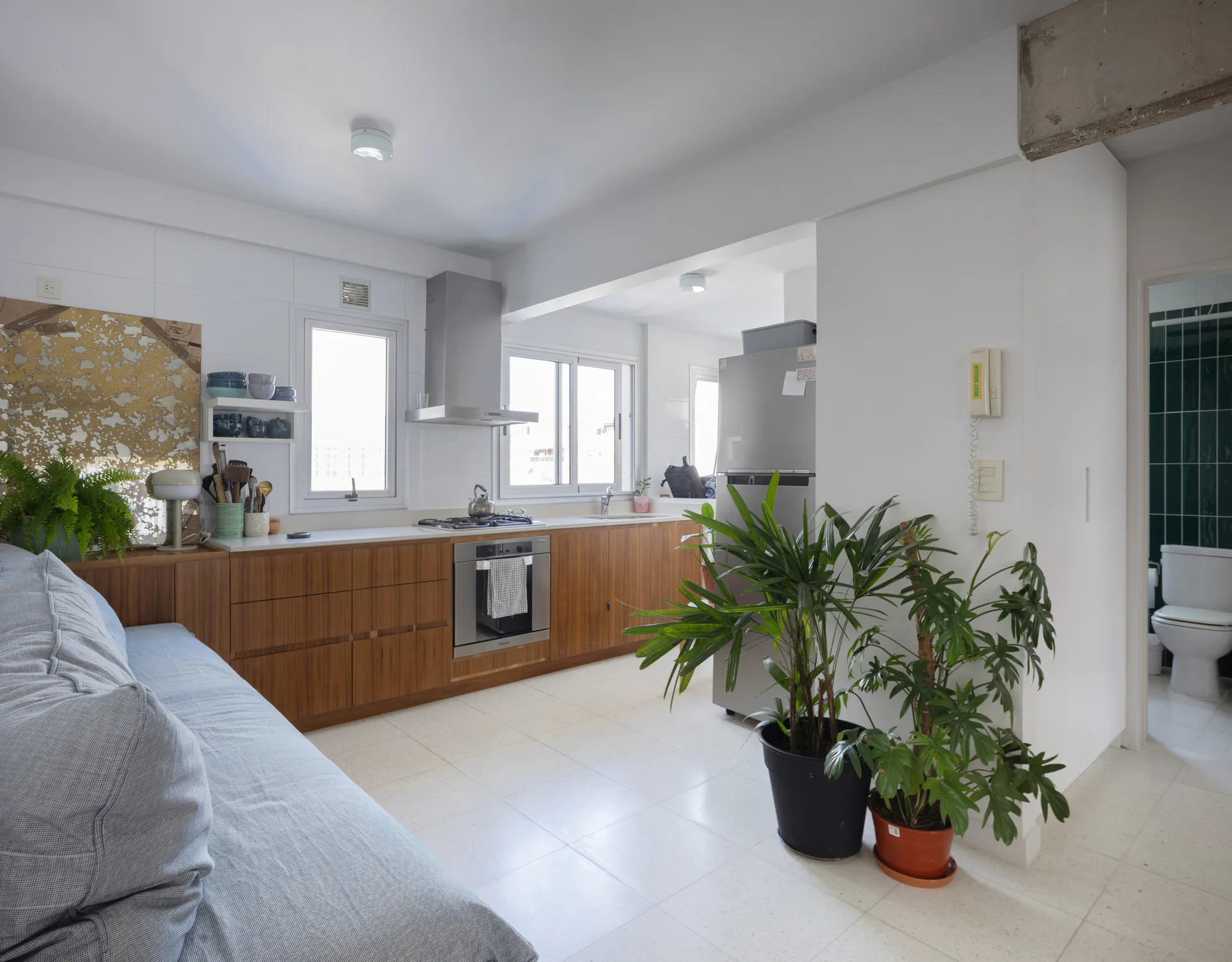 A modern kitchen features medium-toned wood lower cabinets, white walls, large windows, and an exposed concrete beam near a living area with potted plants.