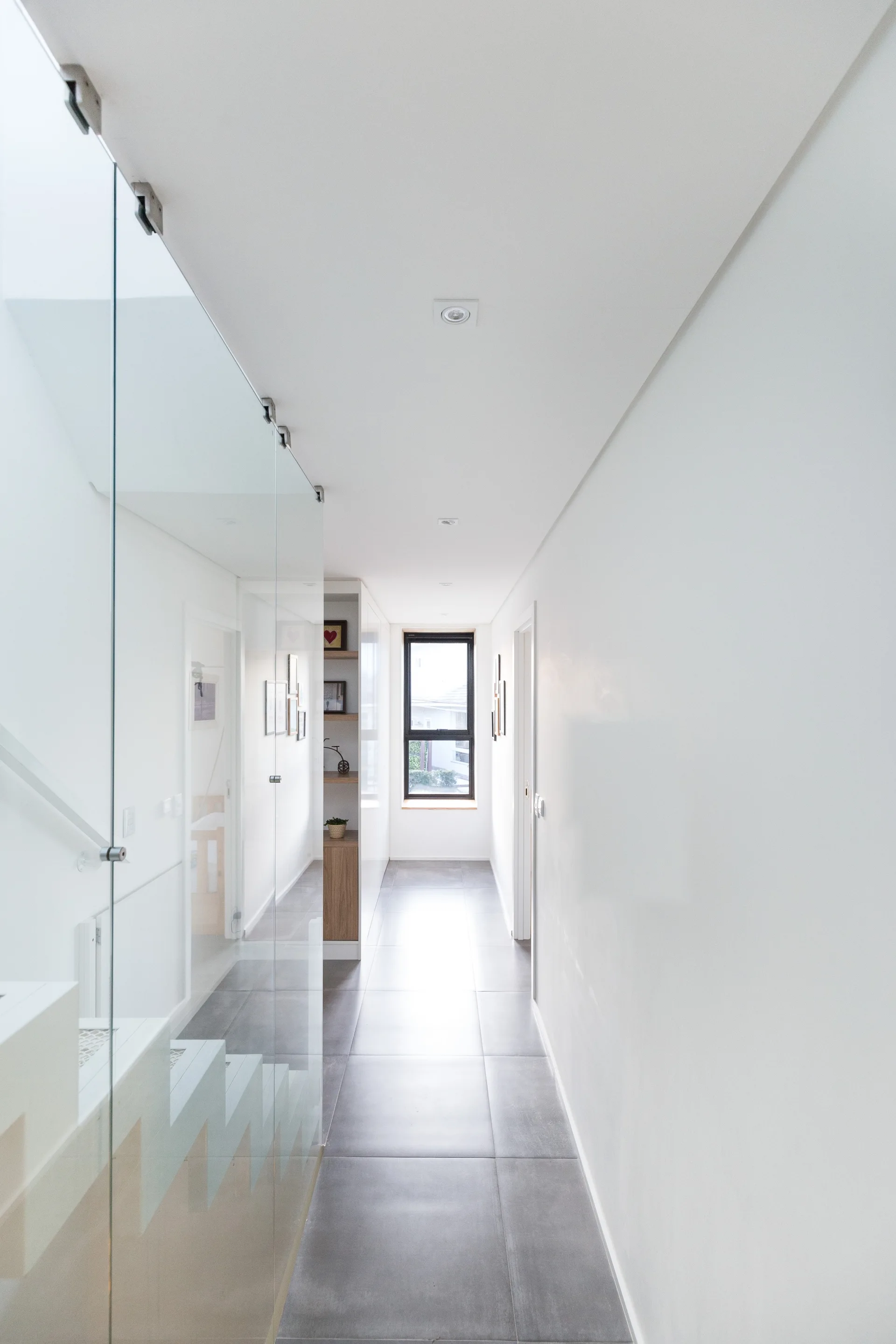 A bright modern hallway features smooth white walls, large grey ceramic floor tiles, a window at the far end, and a full-height glass partition.