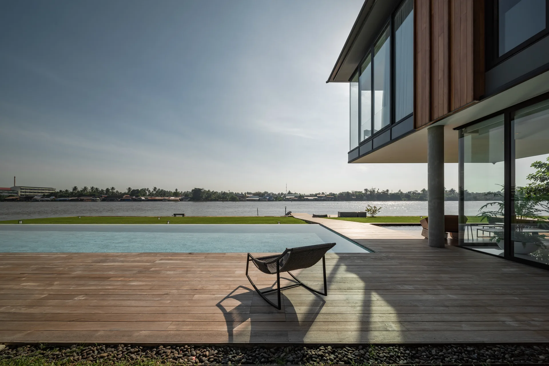 A modern two-story home with wood siding, a wooden deck, and an infinity pool overlooking a wide river with a single chair on the terrace.