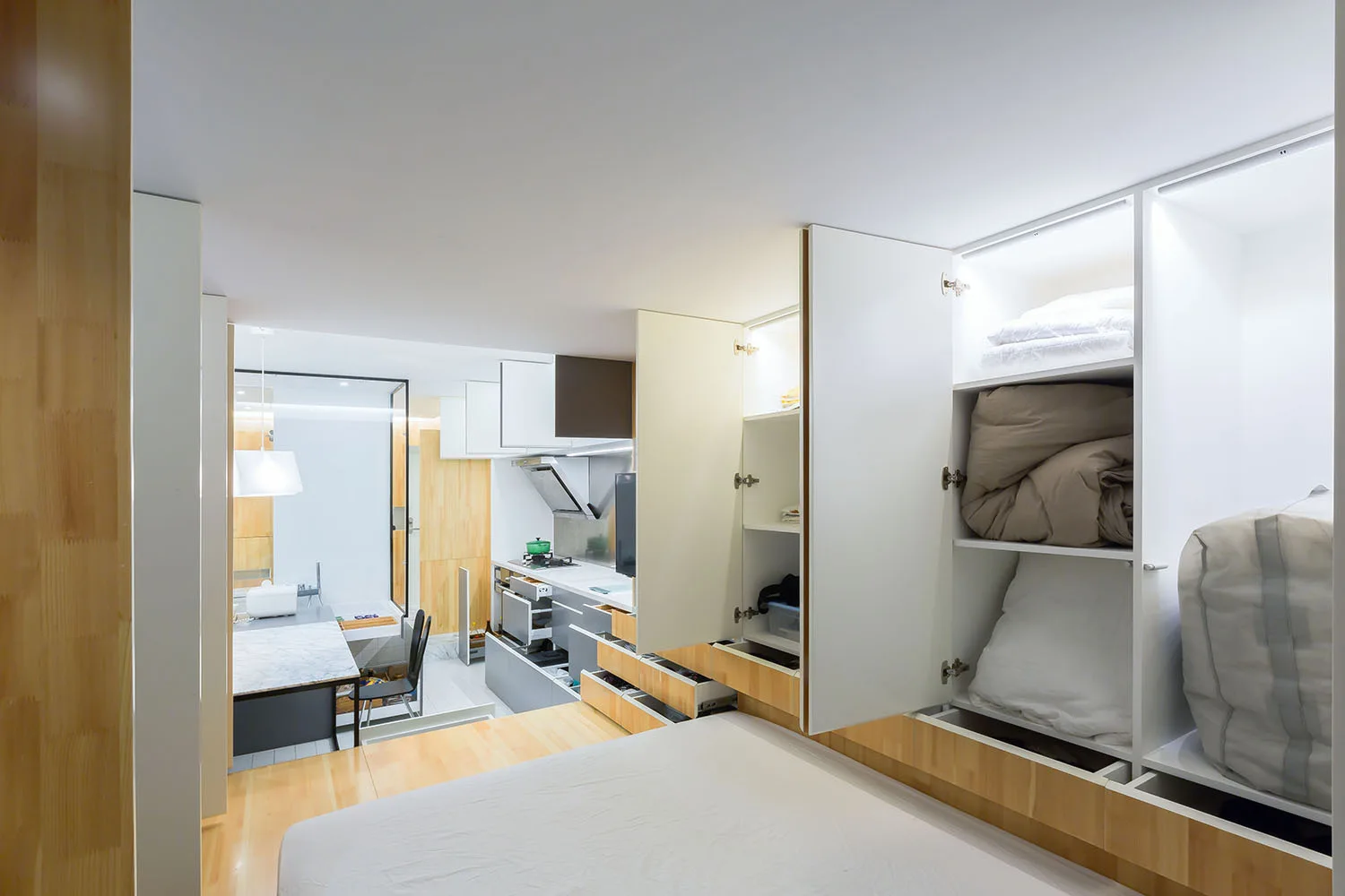 A compact studio apartment showing built-in white cabinets with interior LED lighting next to a bed platform, with a kitchenette in the background.