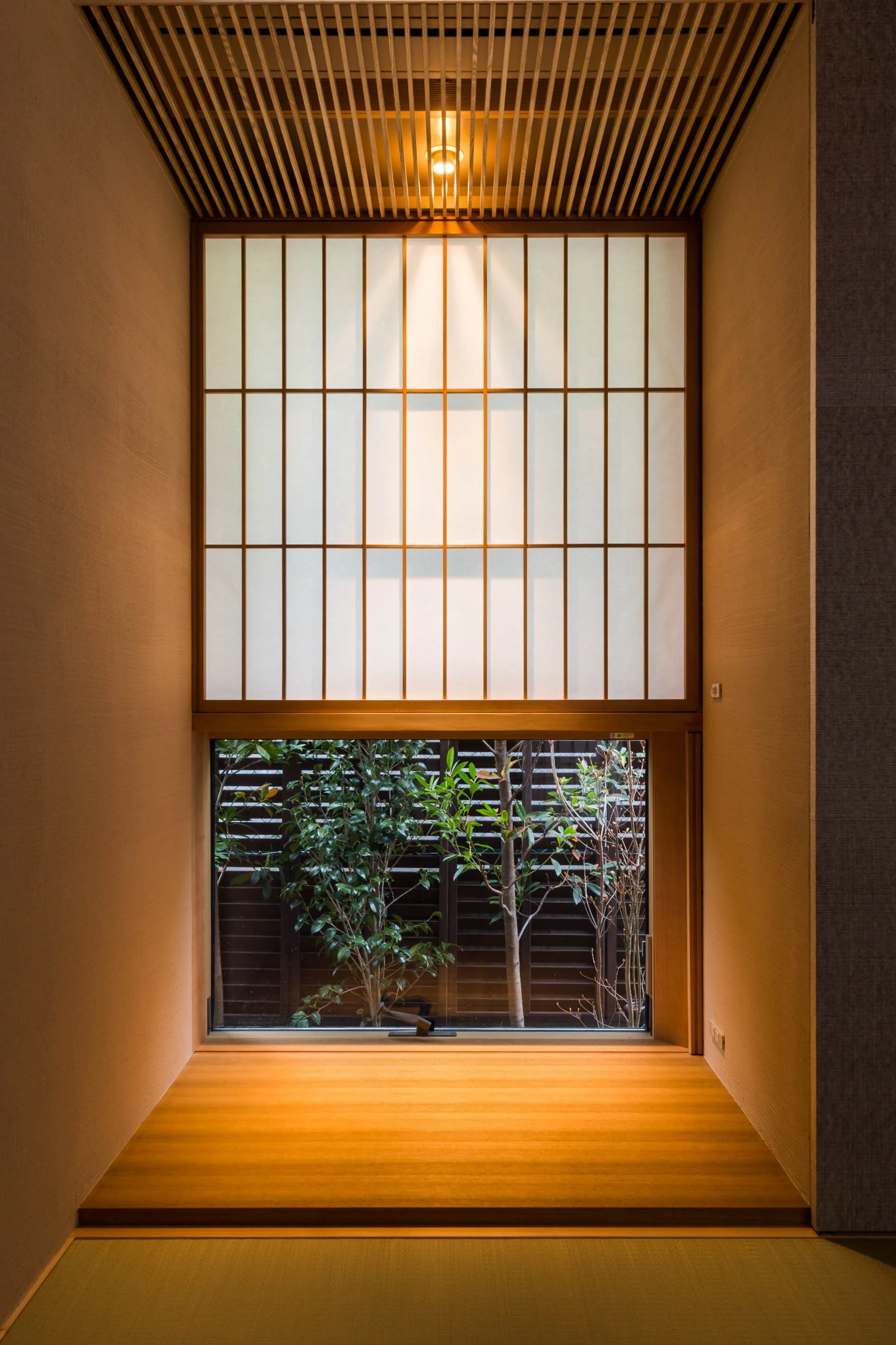 A modern Japanese alcove featuring a slatted wooden ceiling, a raised wood platform, and a shoji screen window framing a view of green plants outside.