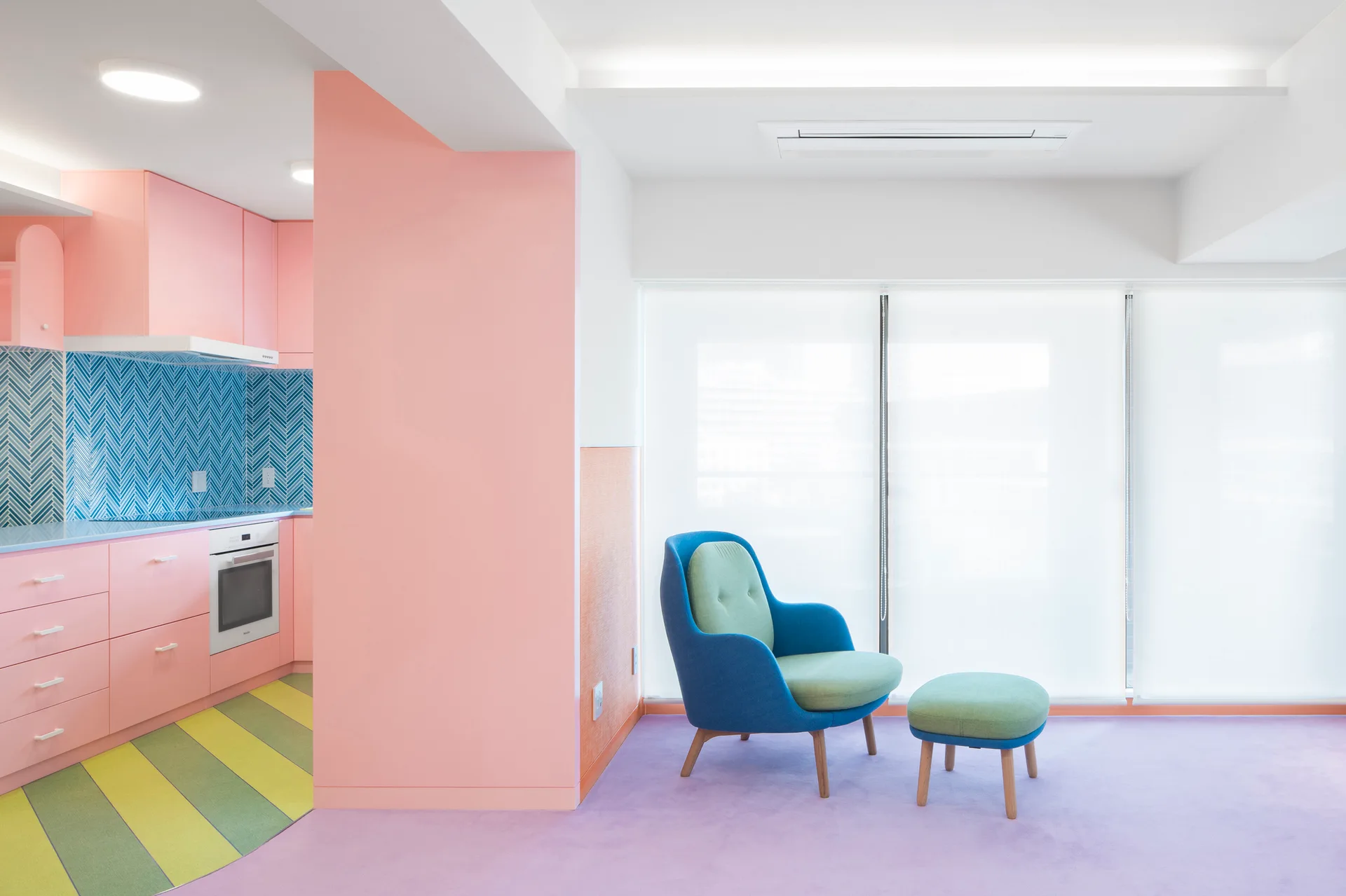 A color-blocked interior featuring pink kitchen cabinets, a blue herringbone backsplash, and a blue armchair on a lavender carpeted floor.