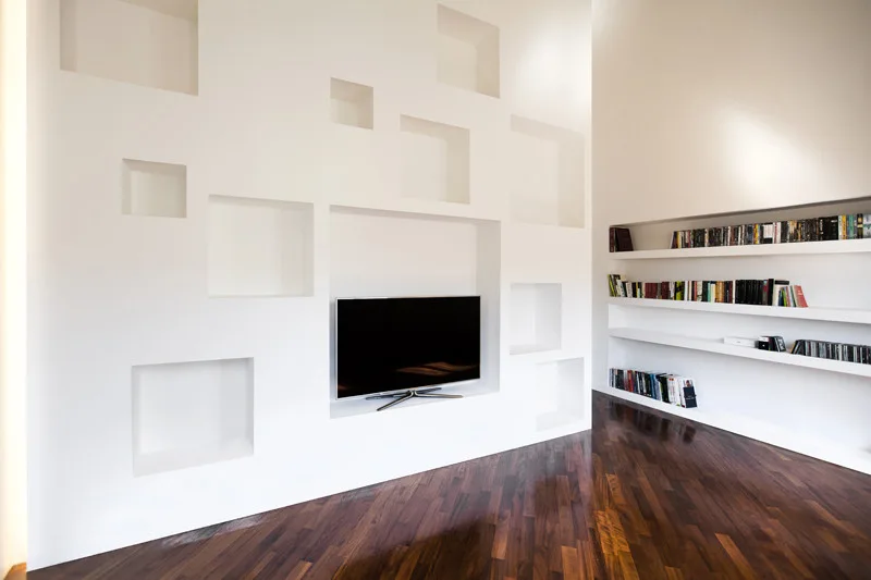 A minimalist white wall features multiple asymmetrical recessed display niches and a television, set above a dark polished chevron wood floor.