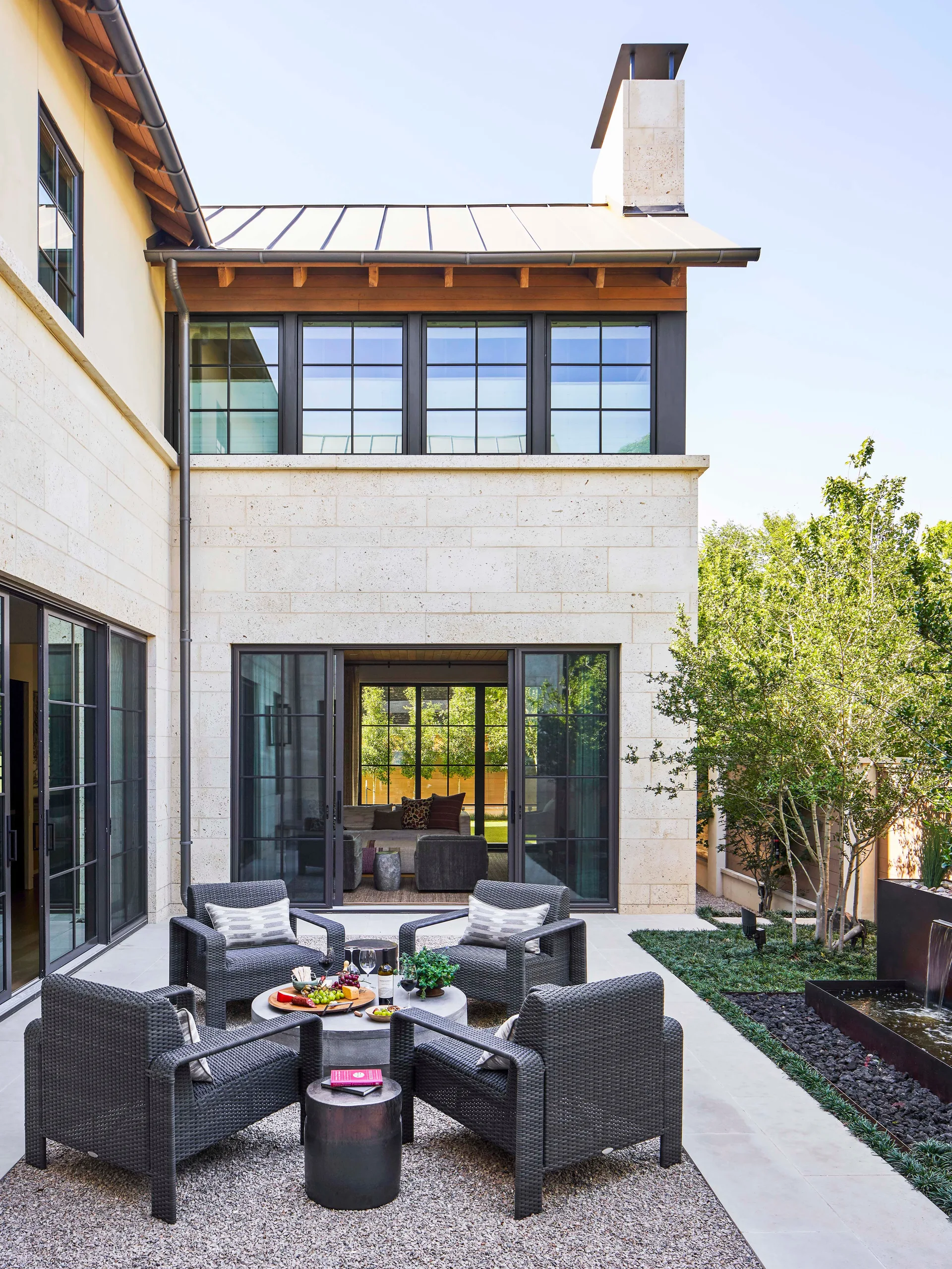 Four dark wicker armchairs sit around a circular table on a gravel patio beside a limestone house with black-framed windows.