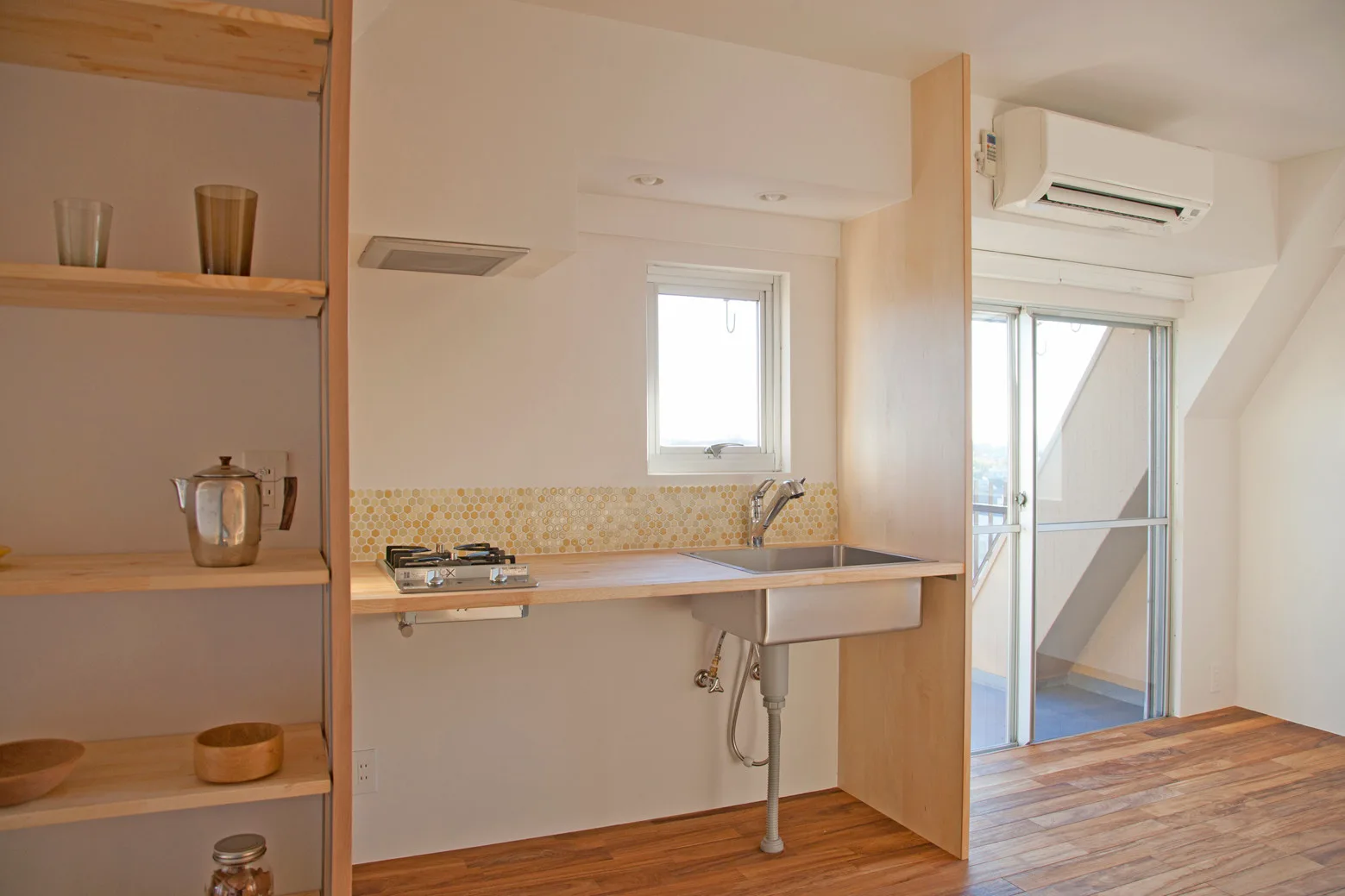 A minimalist kitchenette featuring a light wood countertop, yellow hexagon tile backsplash, stainless steel sink, and open wooden shelving.
