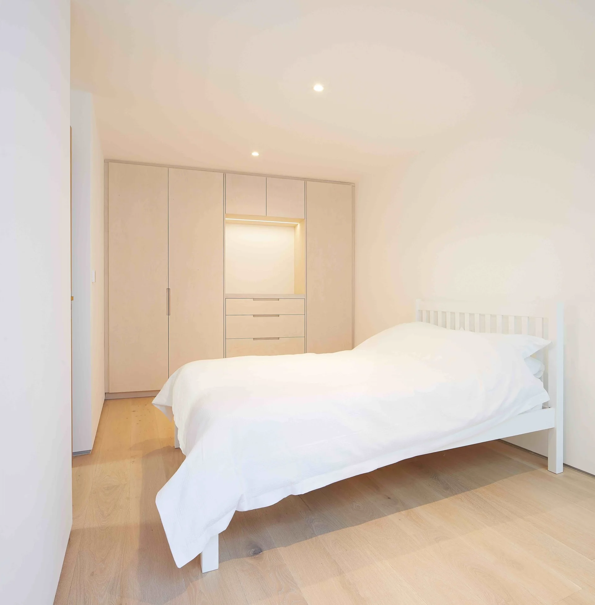 A minimalist bedroom featuring a white wooden bed with white linens, pale wood floors, and a floor-to-ceiling built-in wooden wardrobe with a lit niche.