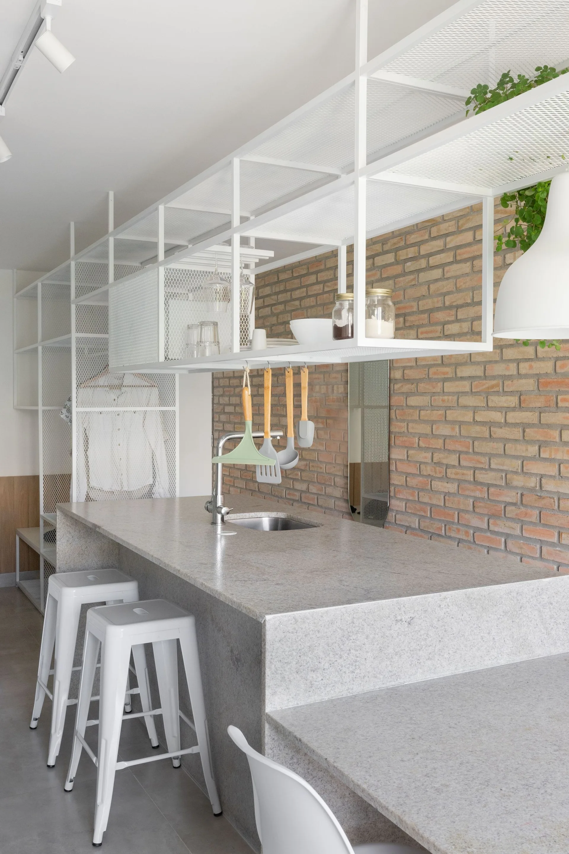 A kitchen island with a grey stone countertop, white metal bar stools, and a white wire mesh overhead shelving unit against an exposed brick wall.