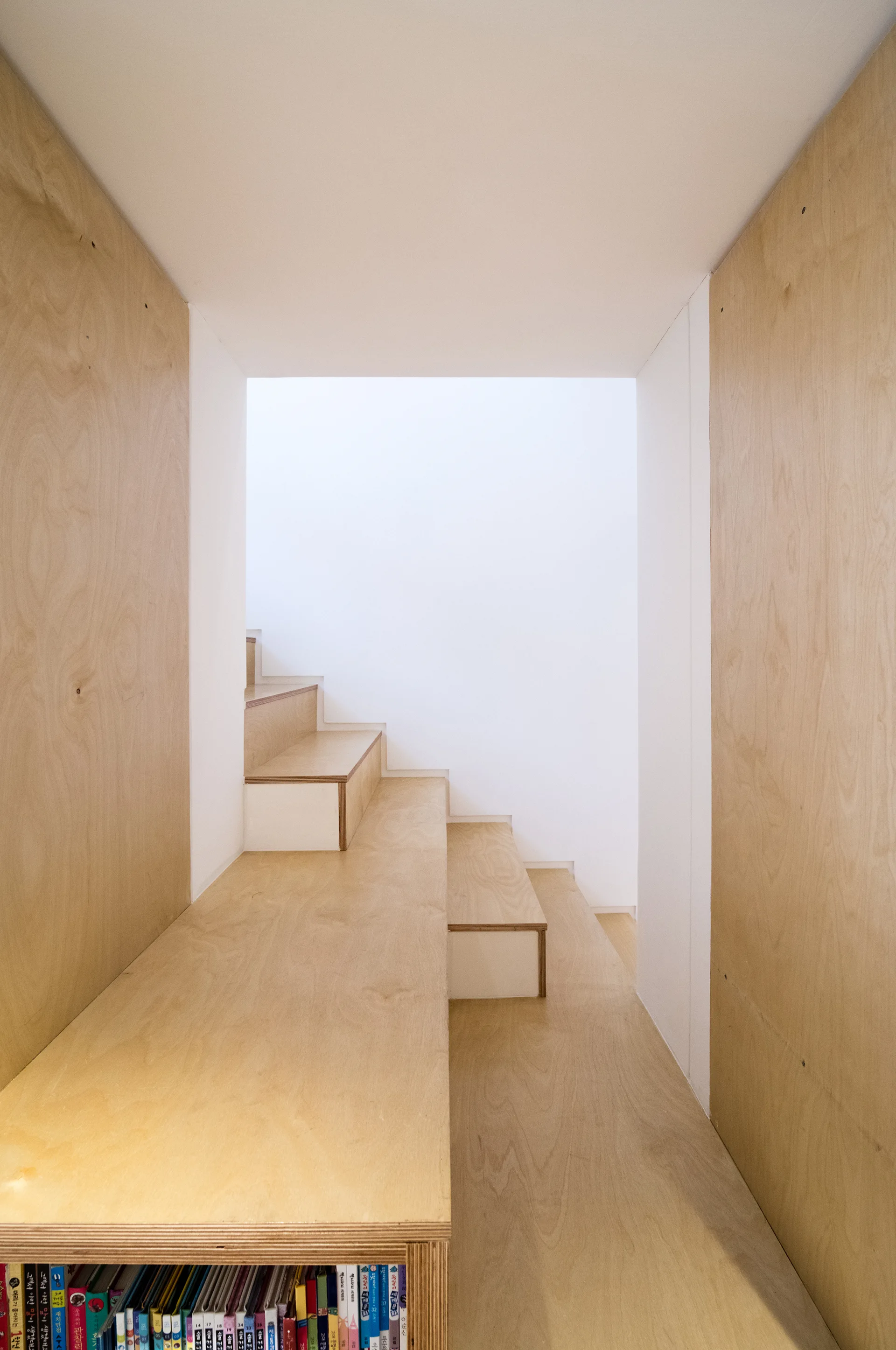 A minimalist plywood staircase with built-in bookshelves at the base, flanked by matching wood paneling and smooth white walls.