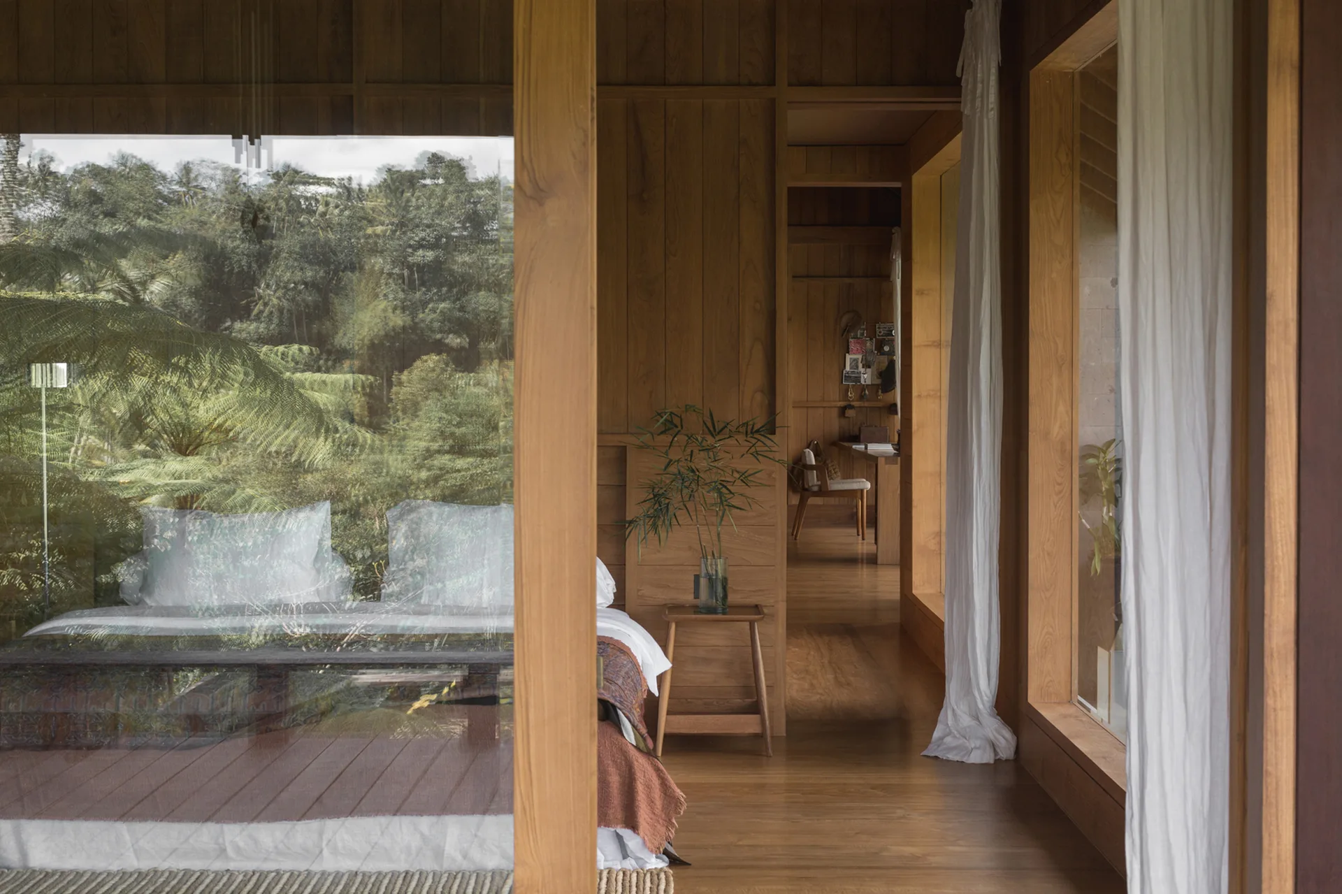 A minimalist wood-paneled bedroom features a bed with white linens and large glass windows reflecting a lush green forest.