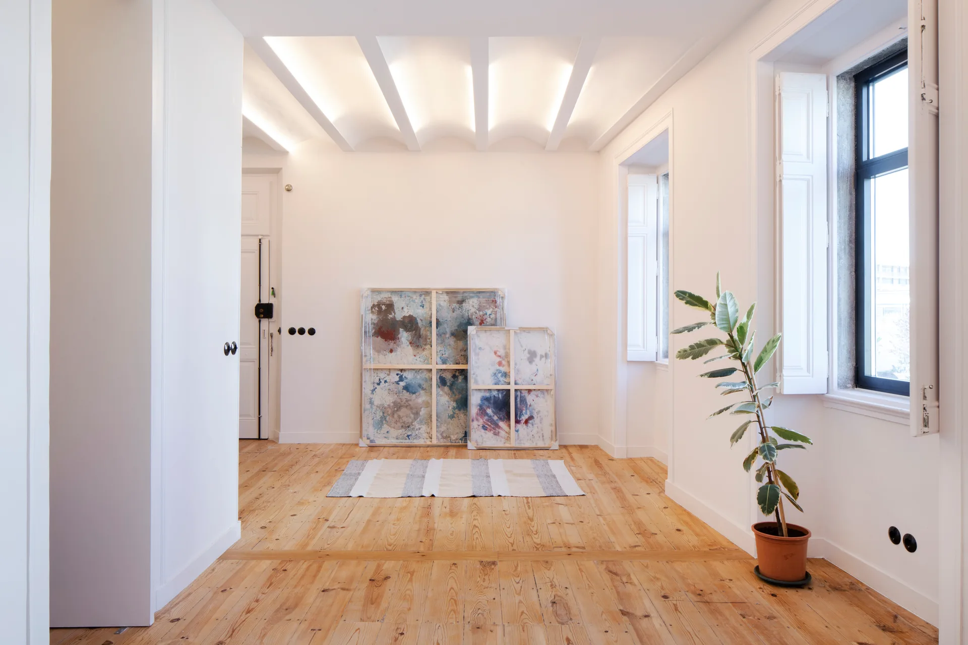 An empty white room features a vaulted cove ceiling with indirect lighting, natural wood floors, shuttered windows, and leaning canvas art.