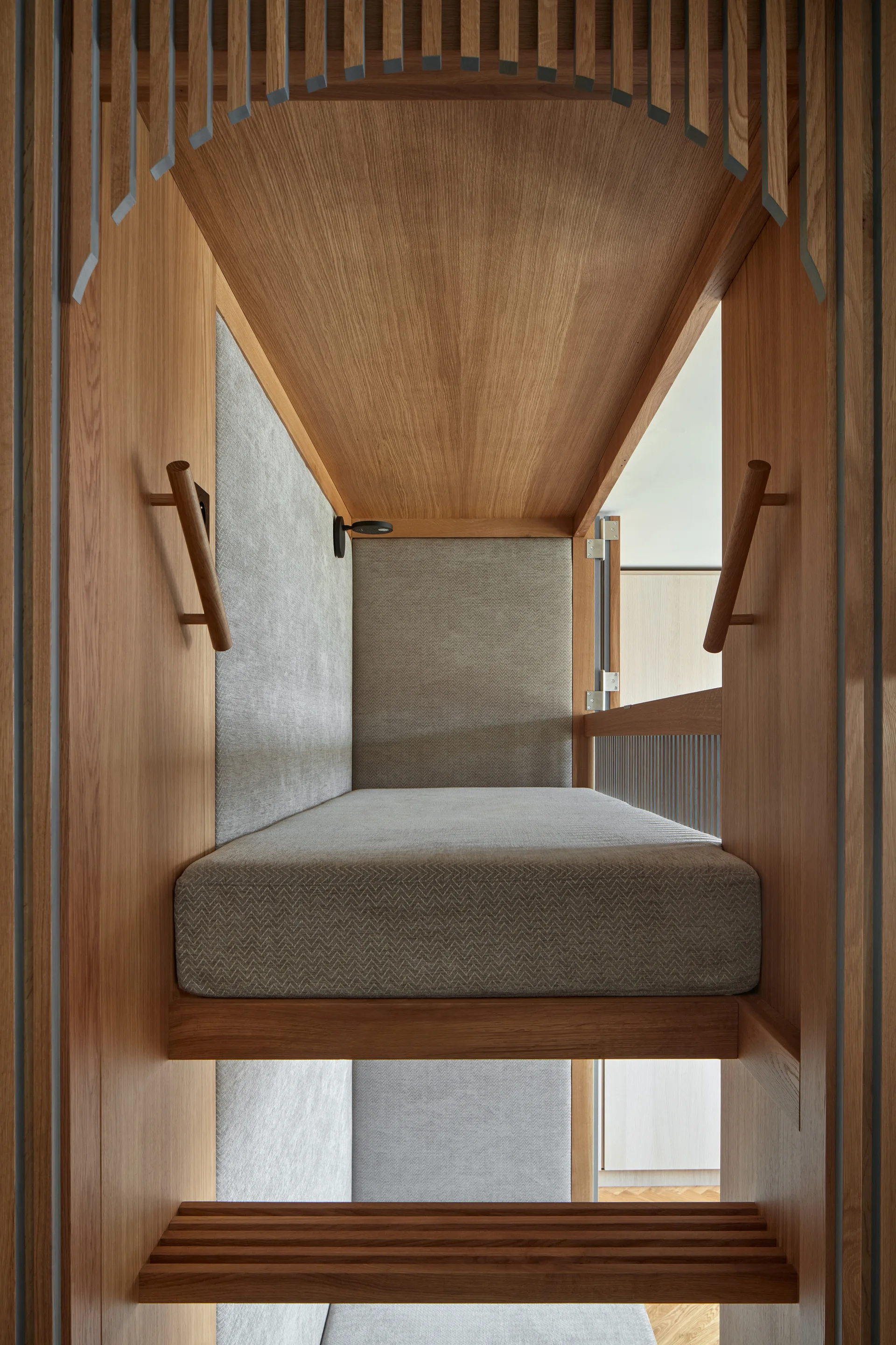 A custom built-in wooden sleeping alcove features grey herringbone upholstery, integrated wooden handles, a wall sconce, and decorative slatted arch details.