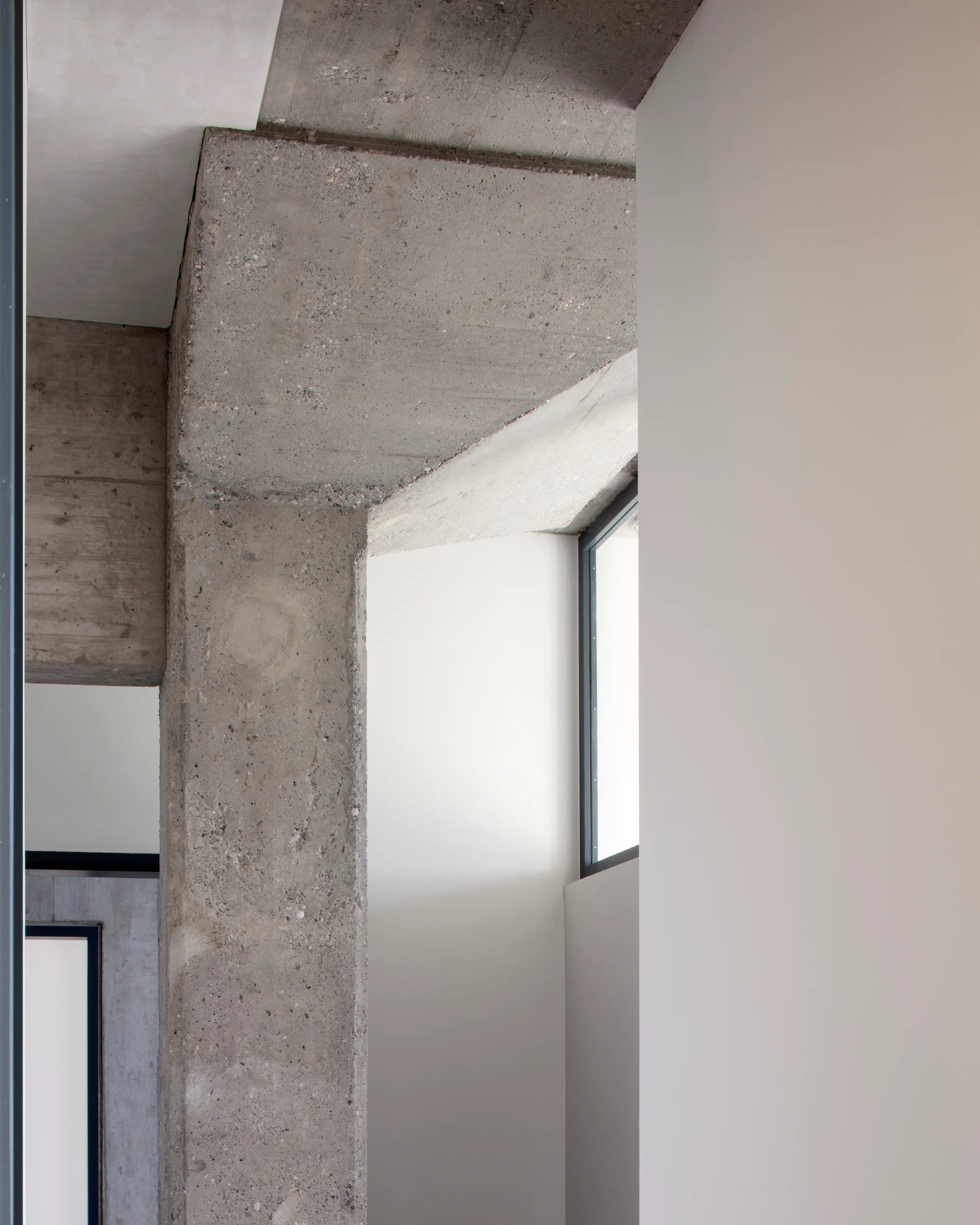 An architectural interior showing a rough grey concrete column and ceiling beam contrasting against smooth painted white plaster walls.
