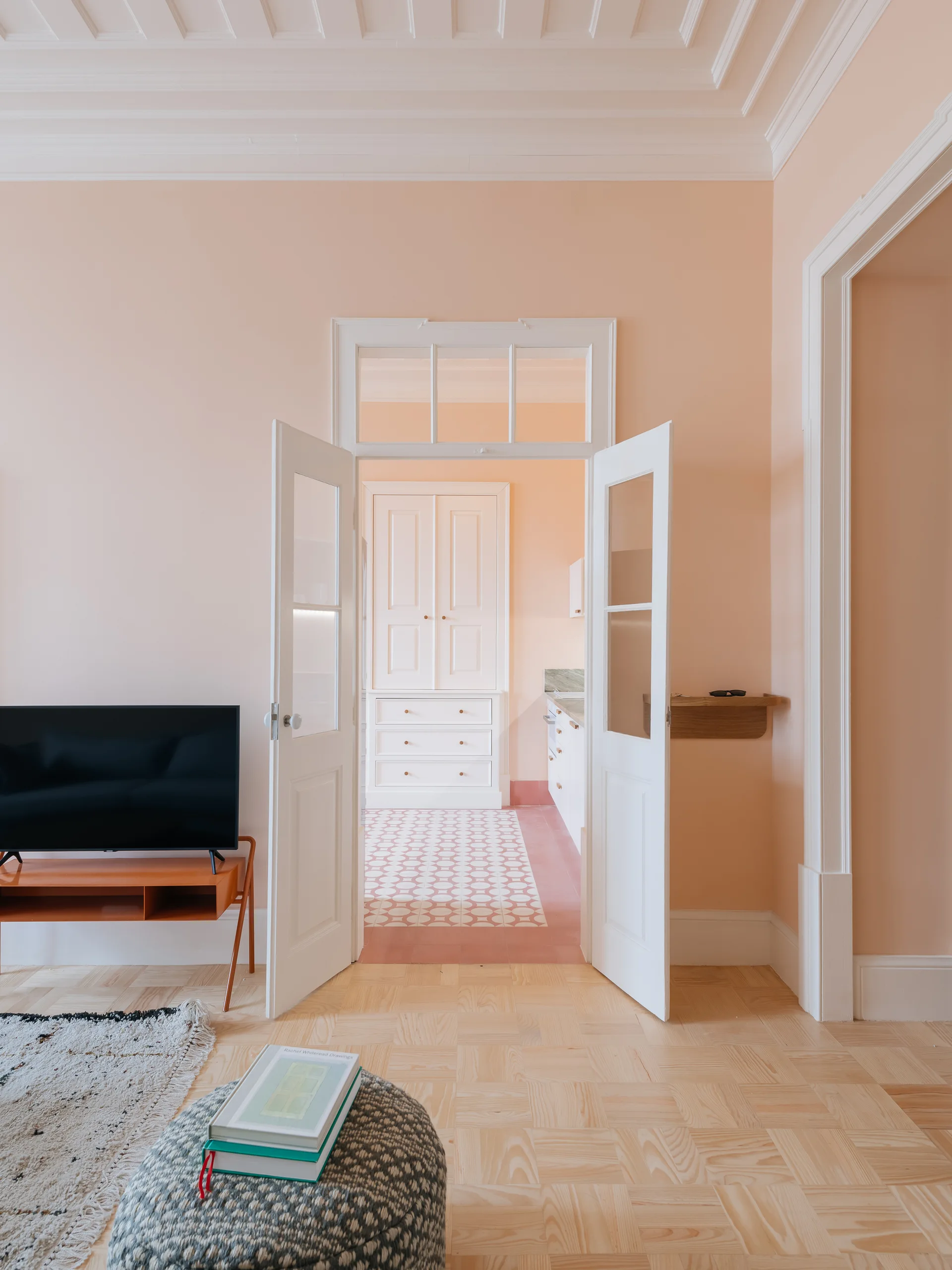An open white double door with a transom window connects a pale pink living room with light parquet floors to a kitchen with patterned tiles.