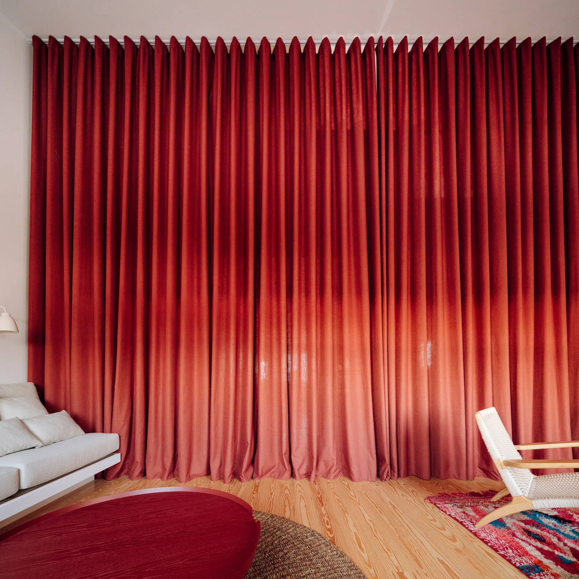 A living room features a massive wall of pleated red curtains, a white sofa, a woven armchair, and light wood plank flooring.