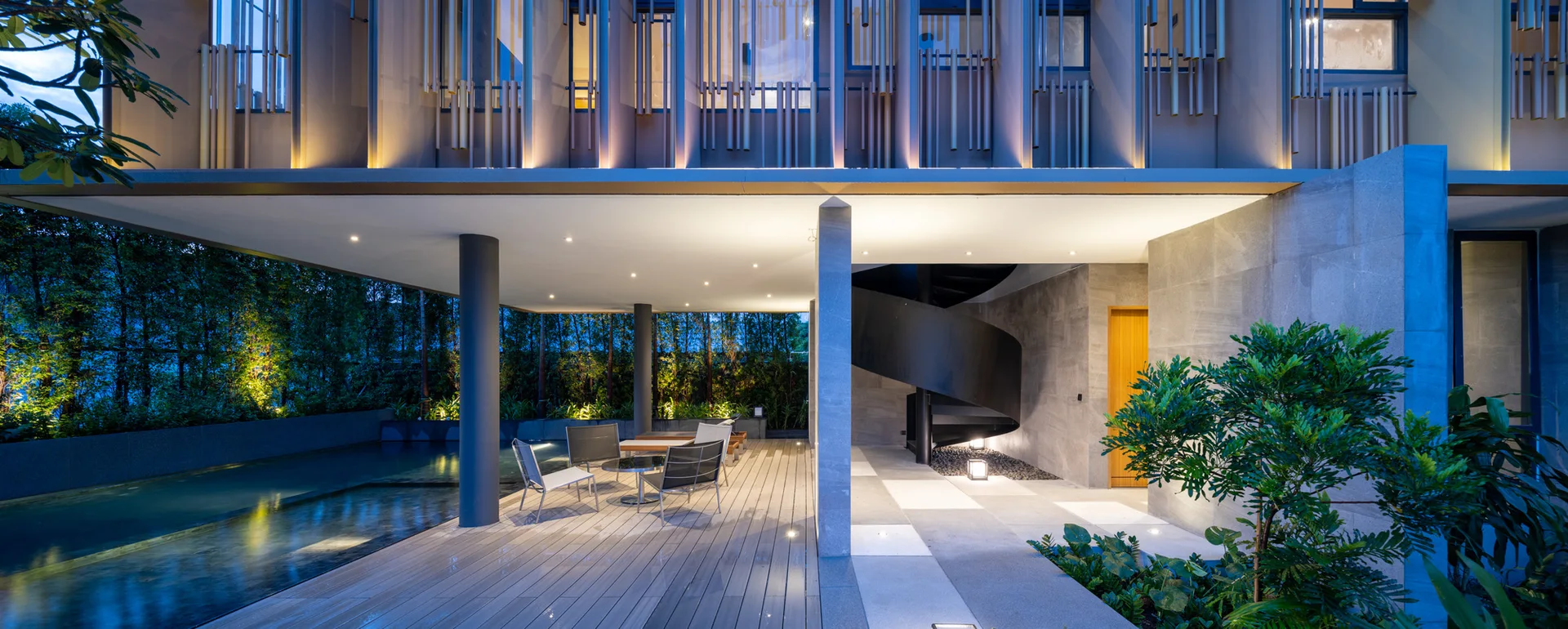 A modern covered patio at dusk featuring a black spiral staircase, stone wall tiling, and an integrated swimming pool.