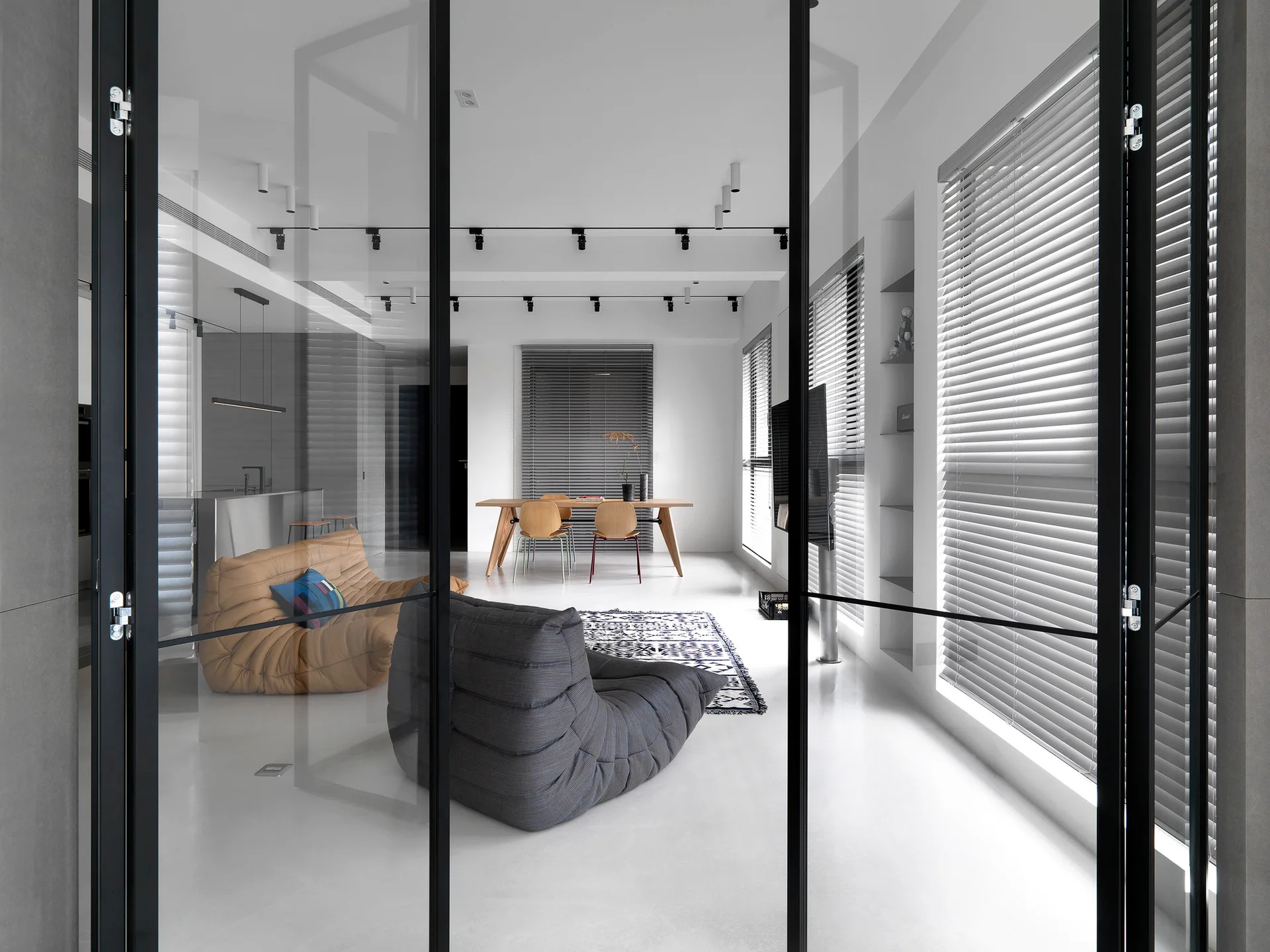 A modern minimalist living room seen through a black metal glass partition door, featuring low-profile sofas and a wooden dining set.