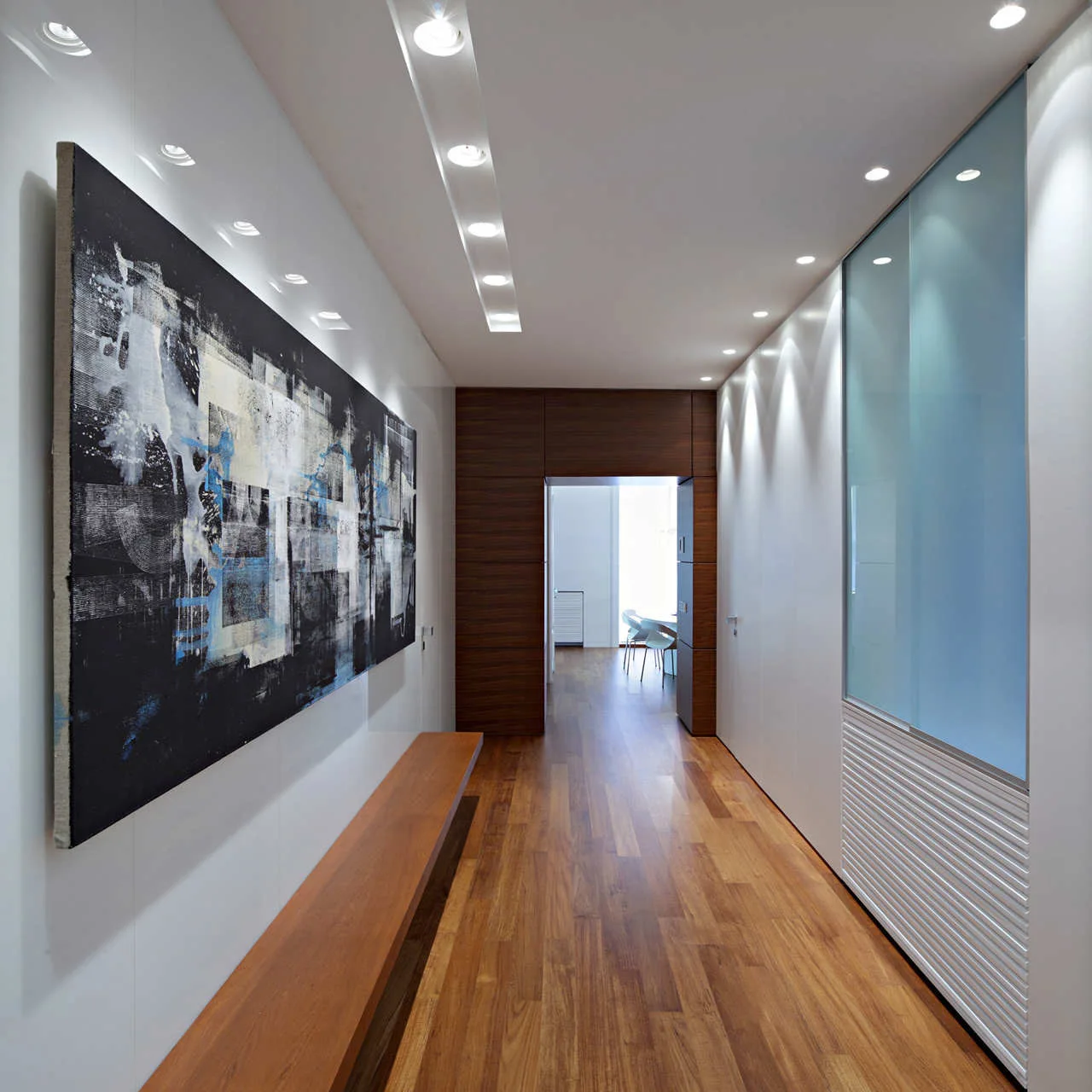 A minimalist white hallway features warm wood flooring, a long wooden bench, a large abstract painting, and recessed ceiling spotlights.