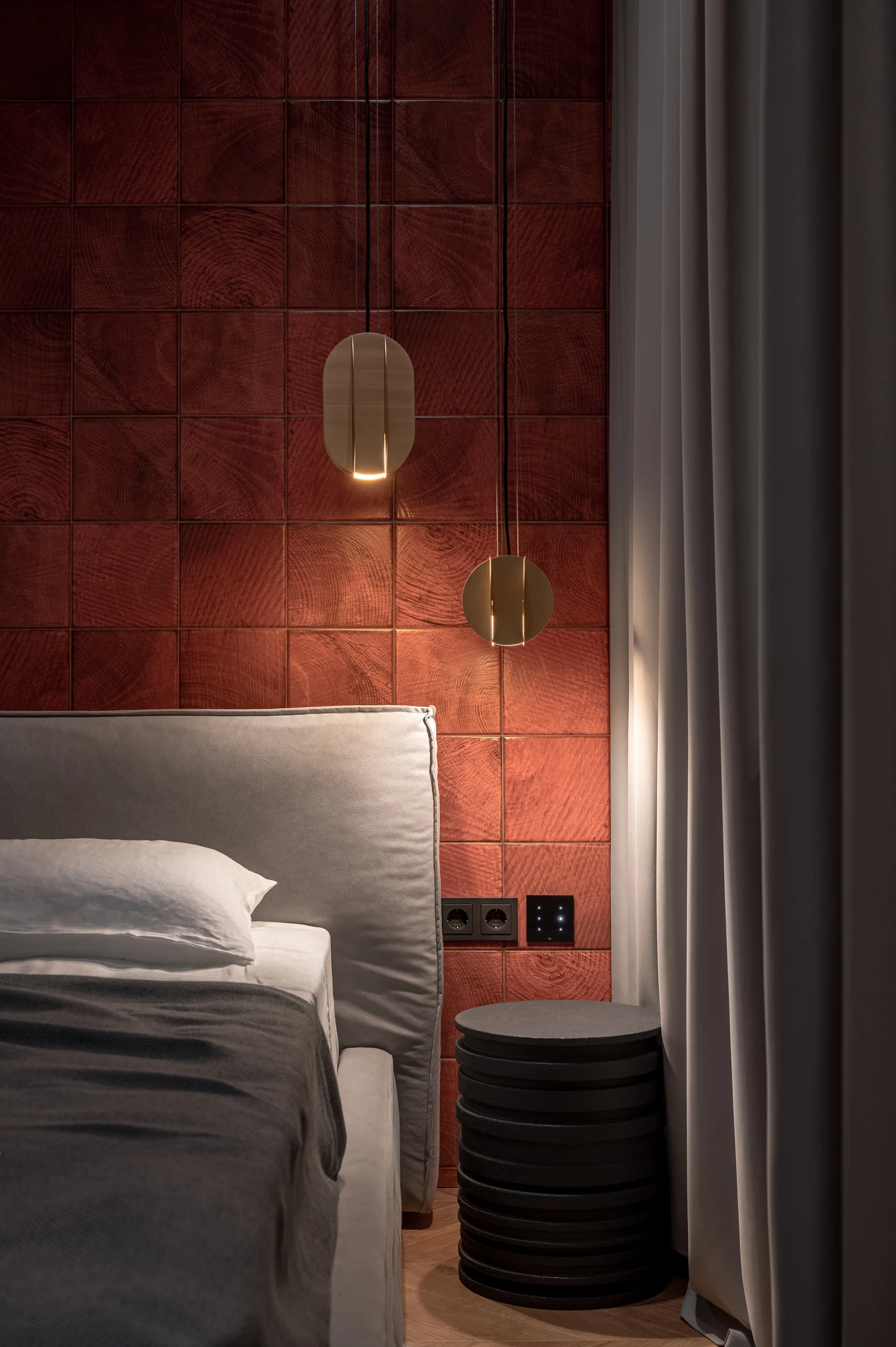 A modern bedroom corner features a bed with a grey upholstered headboard set against a red wood tile accent wall with two warm pendant lights.