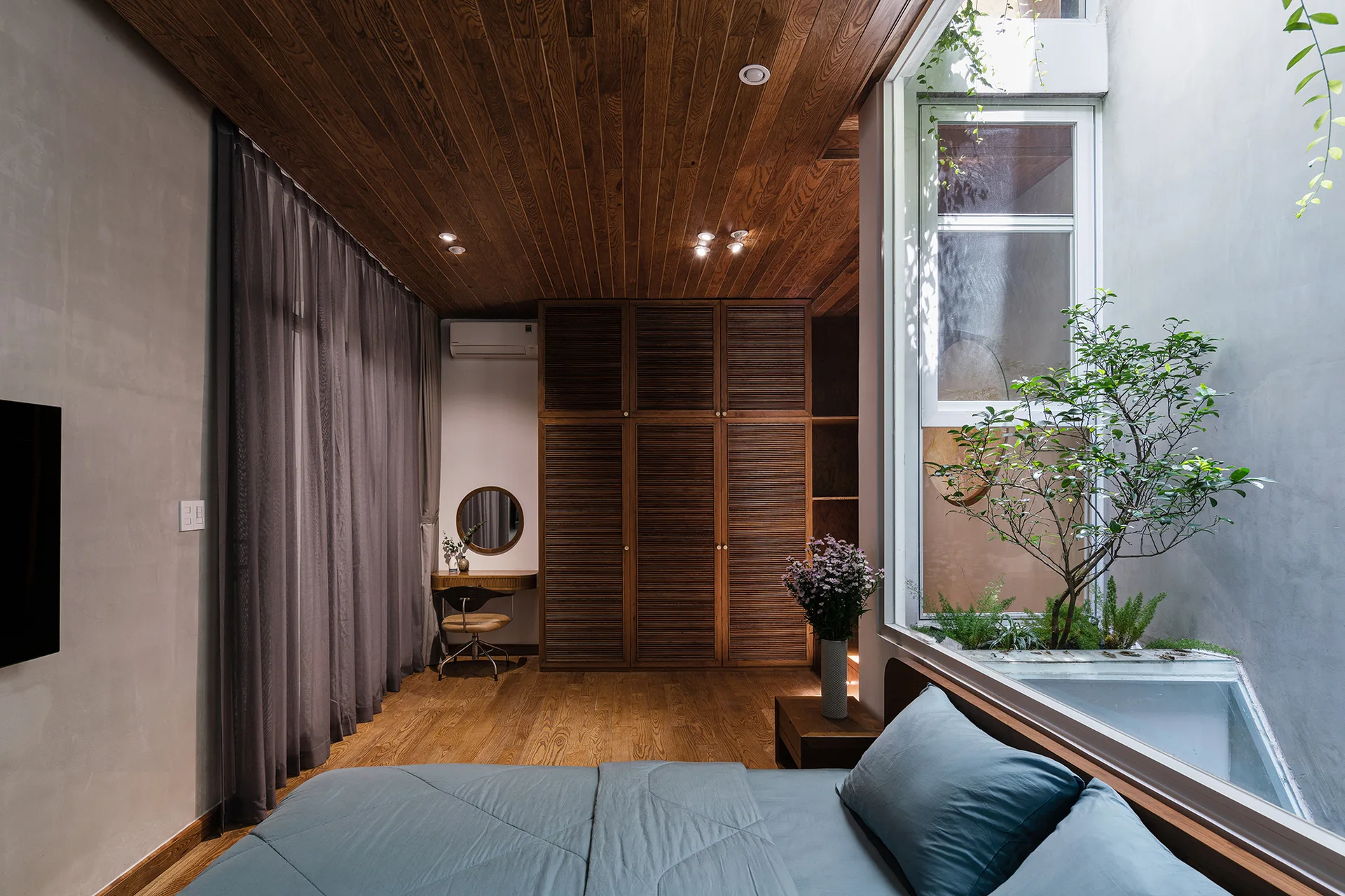 A modern bedroom with wood plank ceilings features a bed with blue sheets and a large window looking into an indoor courtyard garden.