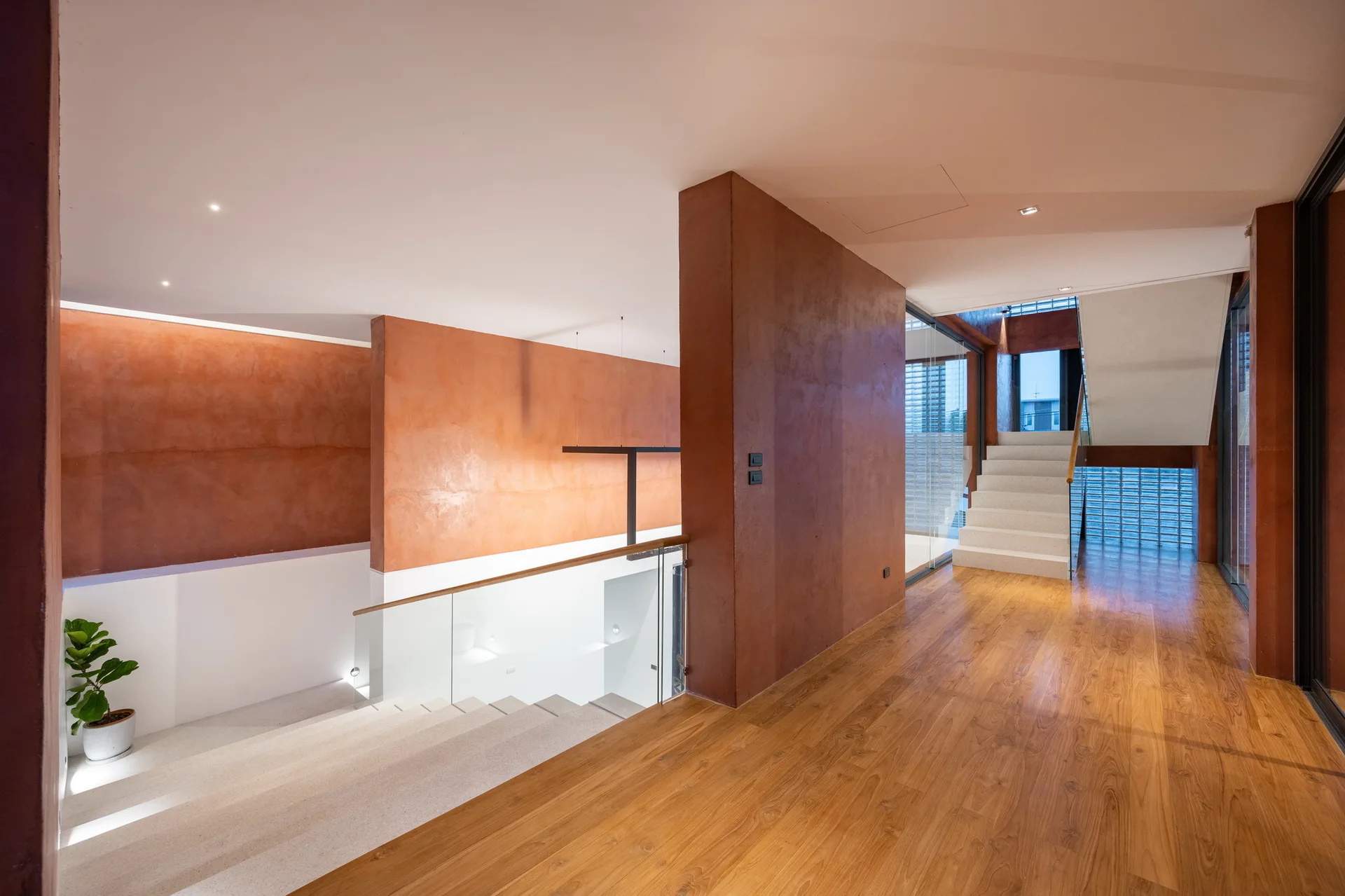 A modern interior hallway features medium wood floors, textured terracotta walls, and a light stone staircase with glass railings.