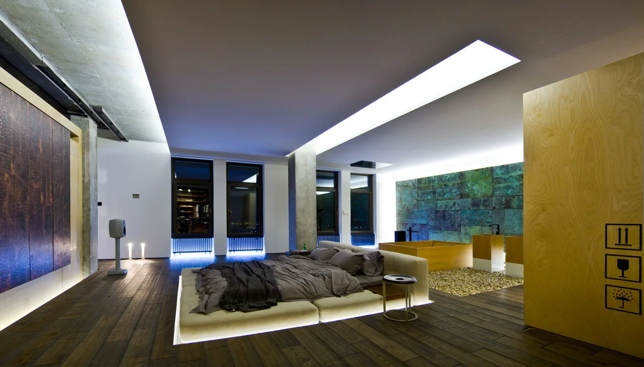 An open-plan loft bedroom featuring a low platform bed with glowing under-bed lighting, concrete pillars, and a wooden soaking tub set on stones.