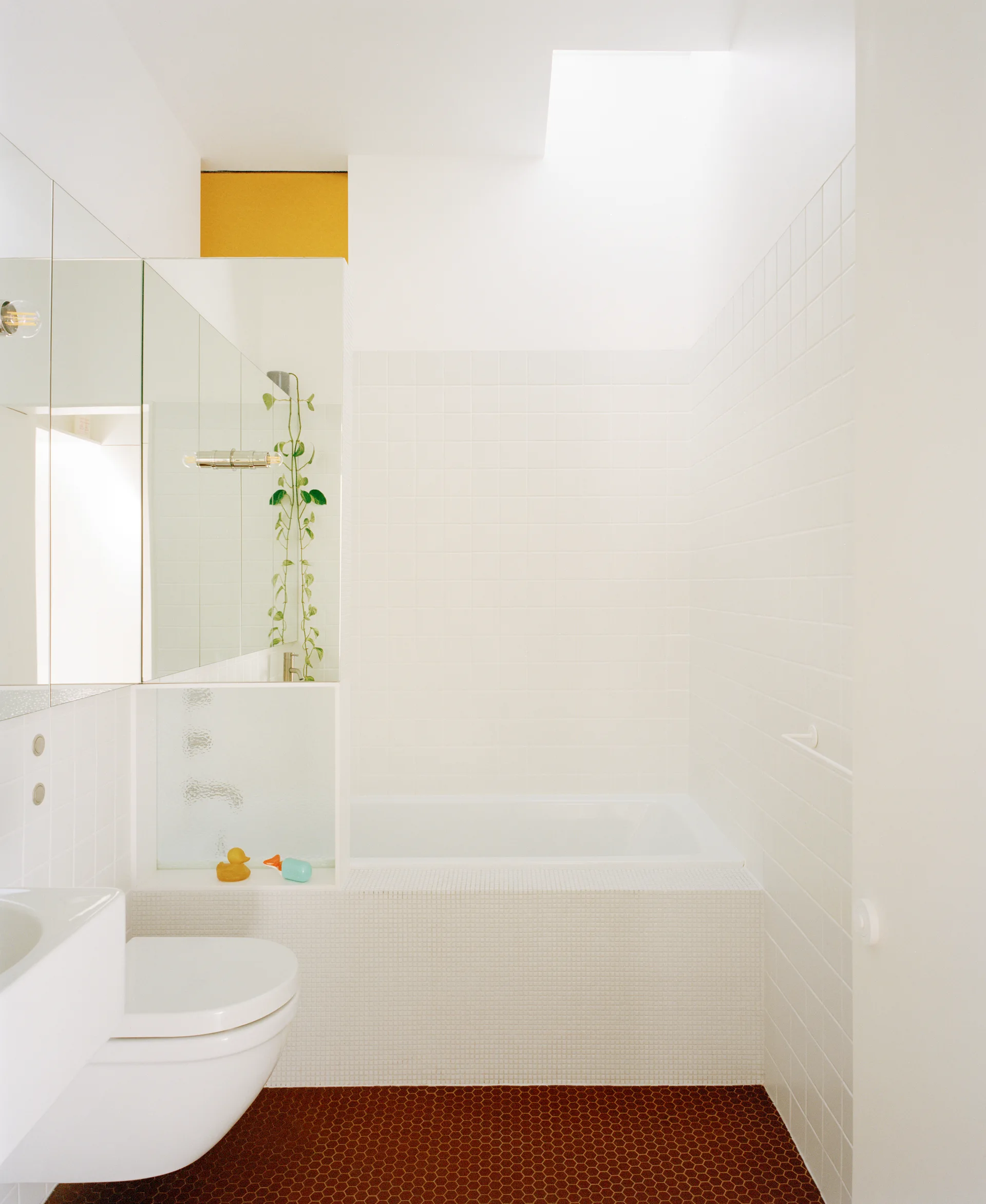 A modern white-tiled bathroom featuring a built-in bathtub under a skylight, a wall-mounted toilet, and terracotta-colored hexagonal floor tiles.