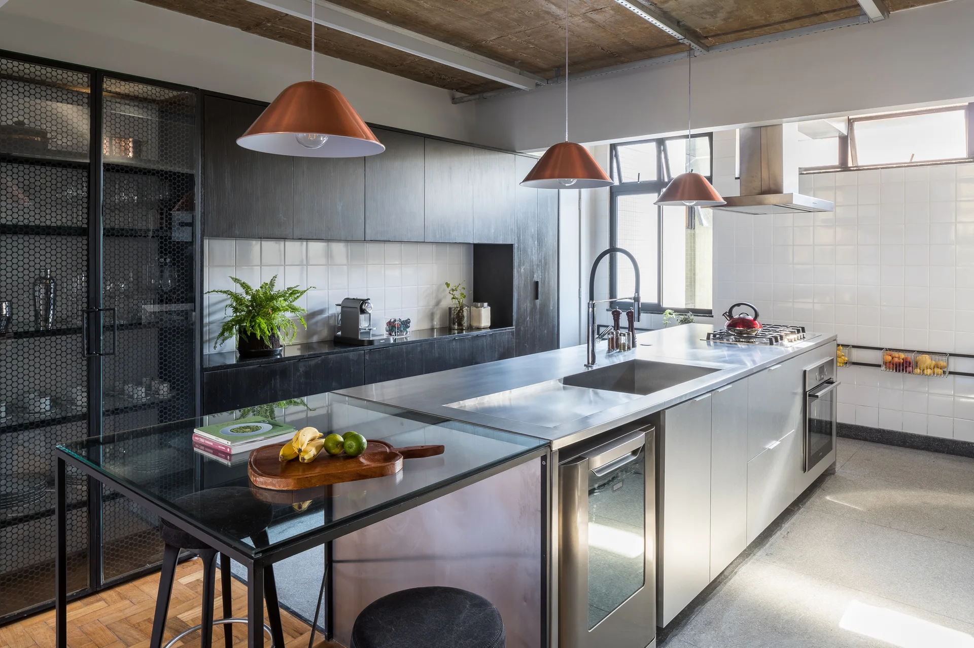 A modern industrial kitchen features a stainless steel island with a glass extension, dark cabinetry, and hanging copper pendant lights.