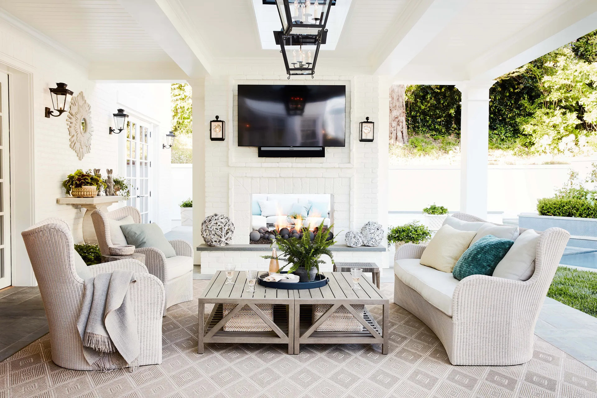 A covered outdoor patio featuring white wicker sofas and armchairs arranged around a square wooden coffee table and a white brick fireplace.