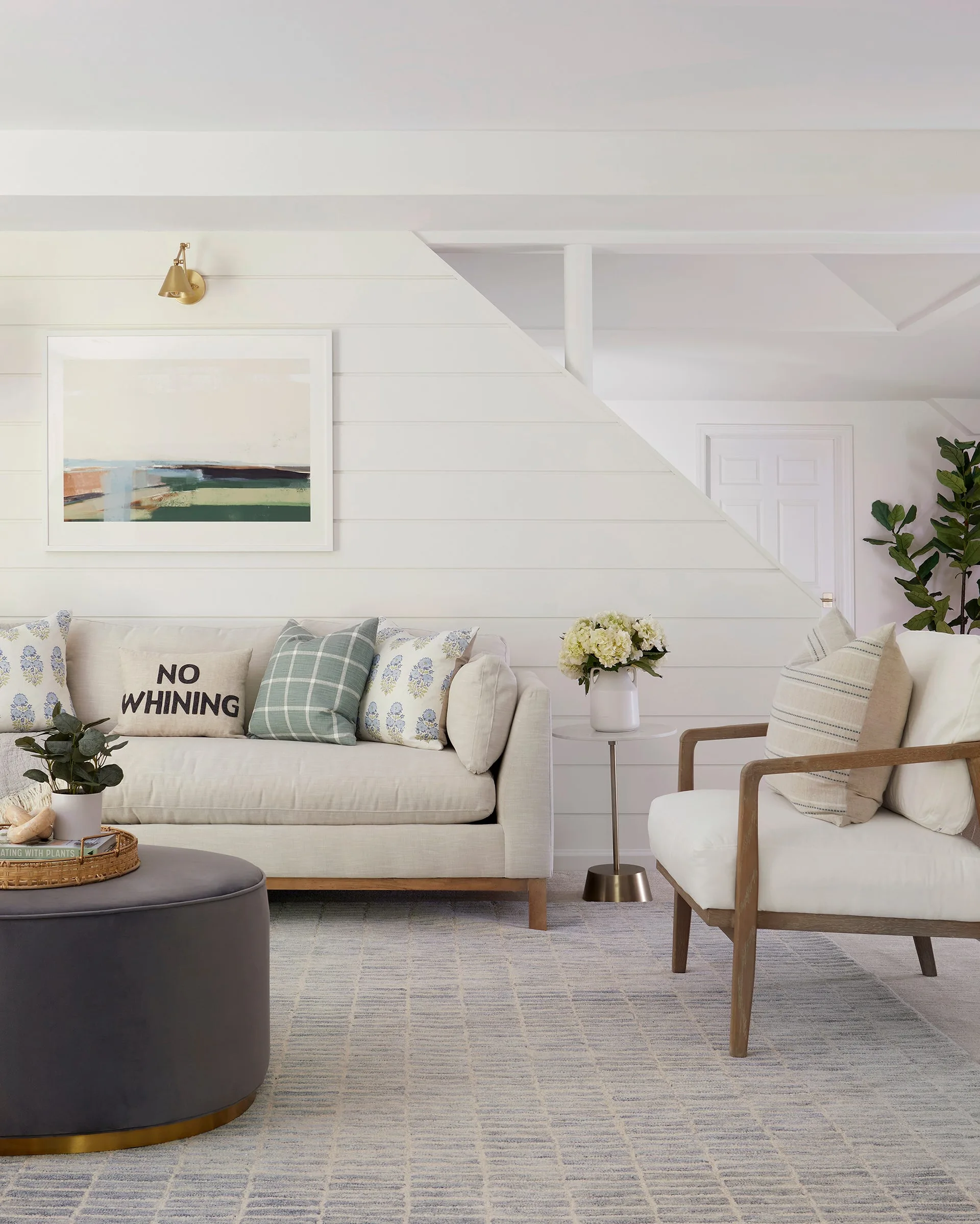 A light living room features a cream sofa with decorative pillows, a wooden armchair, a grey round ottoman, and a white shiplap wall.