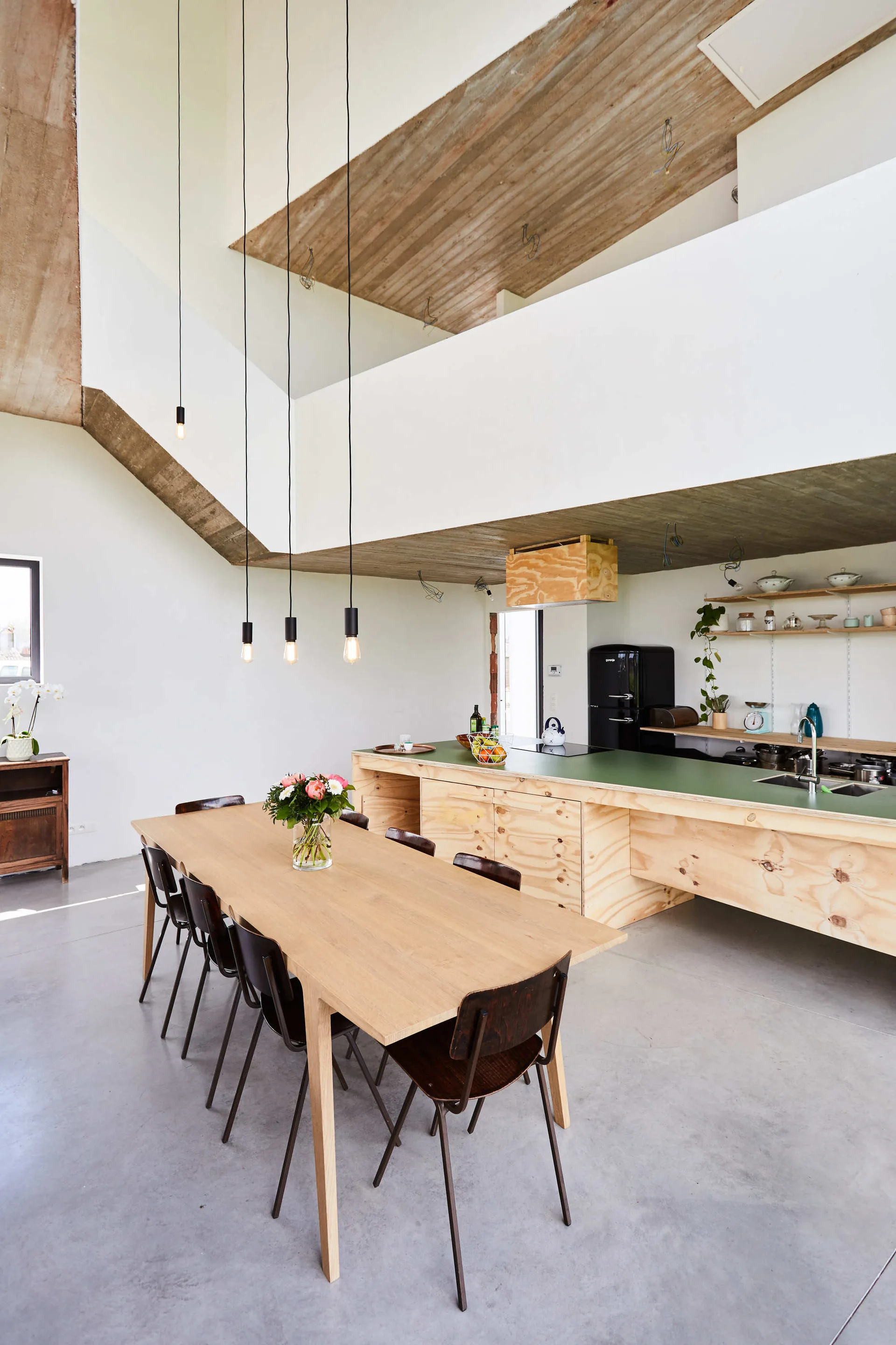 An open-concept kitchen and dining area features a long plywood island with a green countertop, a wooden table, and an exposed concrete ceiling.