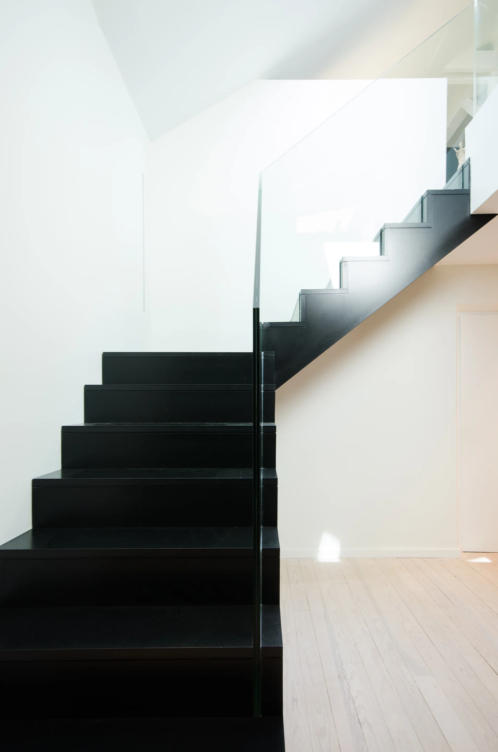 A modern staircase features matte black steps and a clear glass railing set against white walls and light wood flooring.