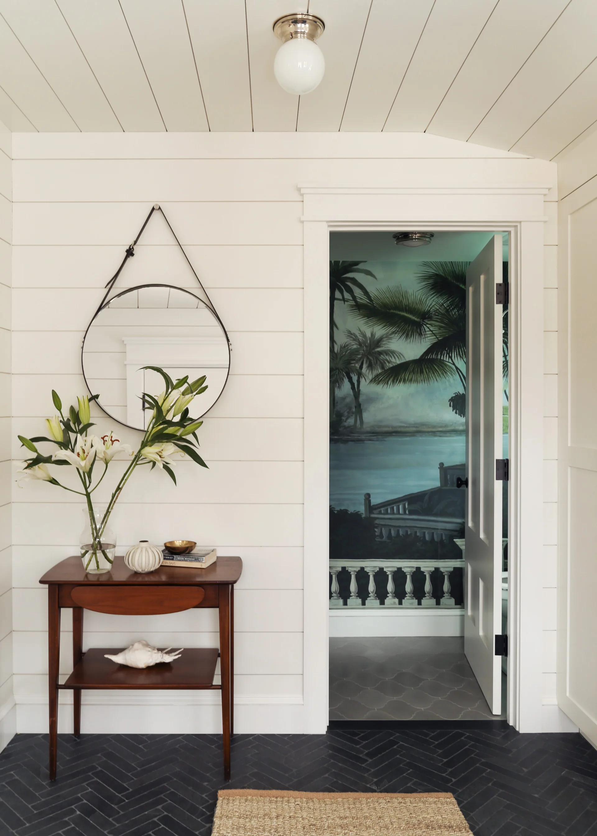 A white shiplap entryway features a wooden console table under a round hanging mirror, with a doorway opening to a room with a tropical mural.