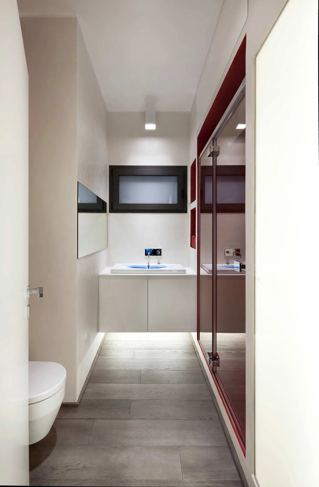 A narrow modern bathroom featuring a floating white vanity with a blue sink basin, grey wood-look plank flooring, and a glass shower door.