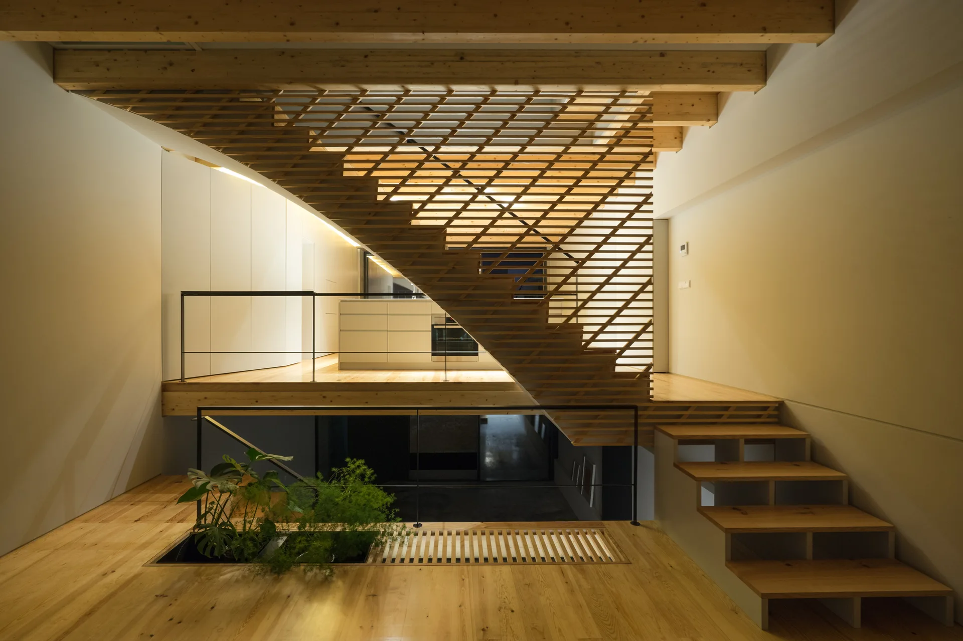 A split-level home interior featuring a central wooden slatted staircase, natural wood floors, exposed ceiling beams, and a built-in floor planter.