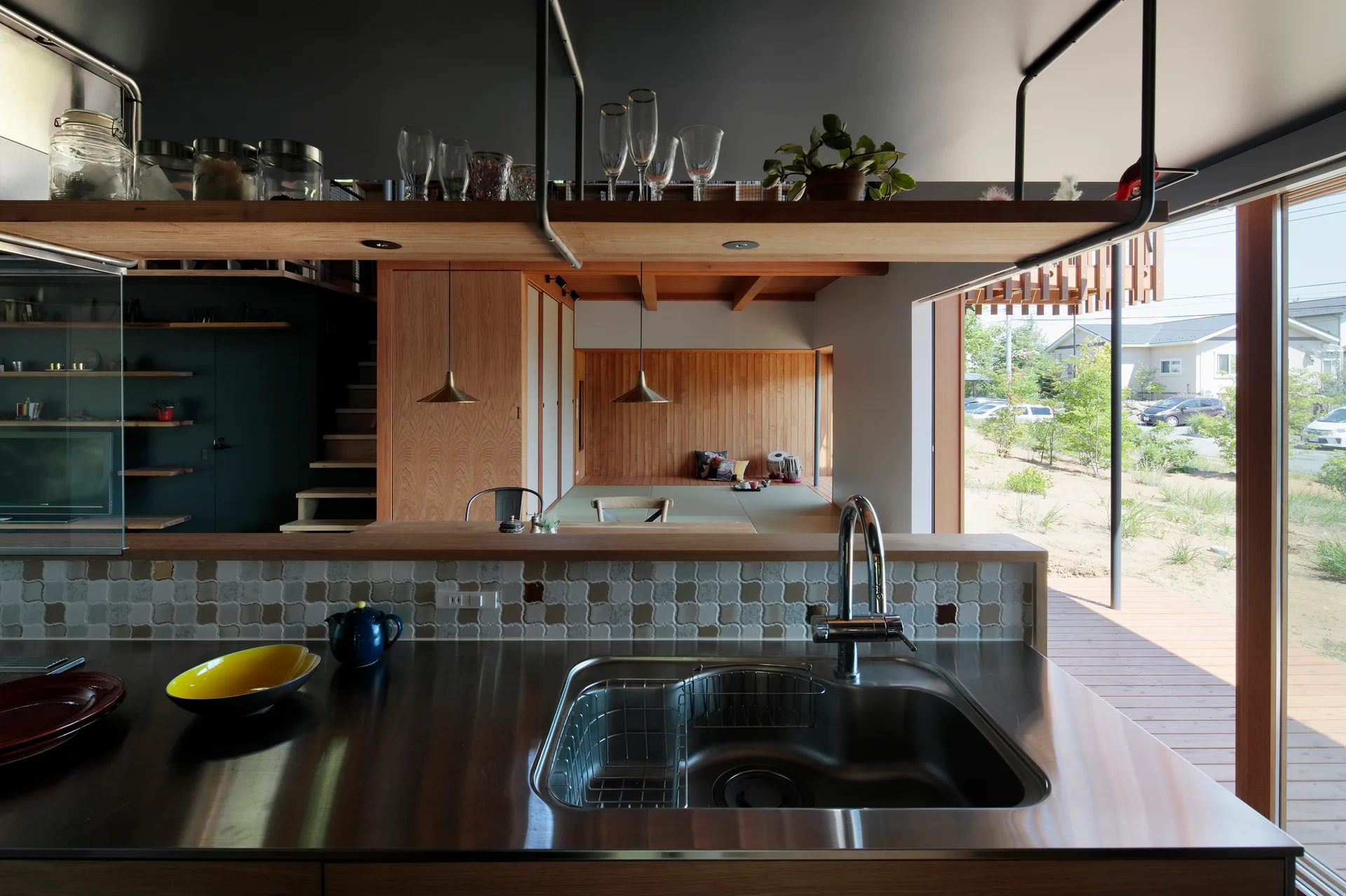 A kitchen with a stainless steel countertop and suspended wood shelf looks out to a wood-paneled living room with sliding glass doors.