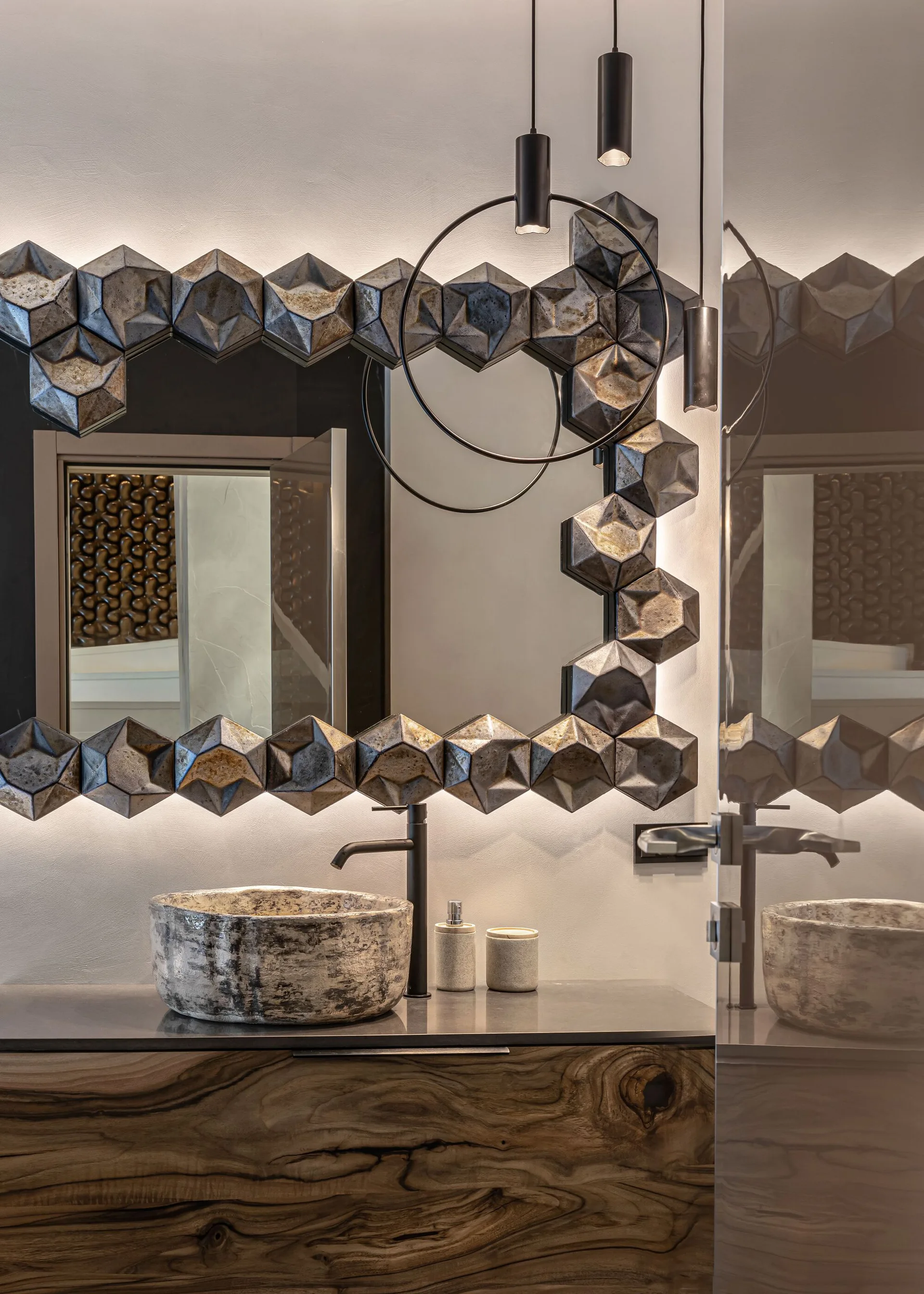 A modern bathroom vanity with a natural wood cabinet, a textured stone vessel sink, and a backlit sculptural mirror with a faceted geometric frame.