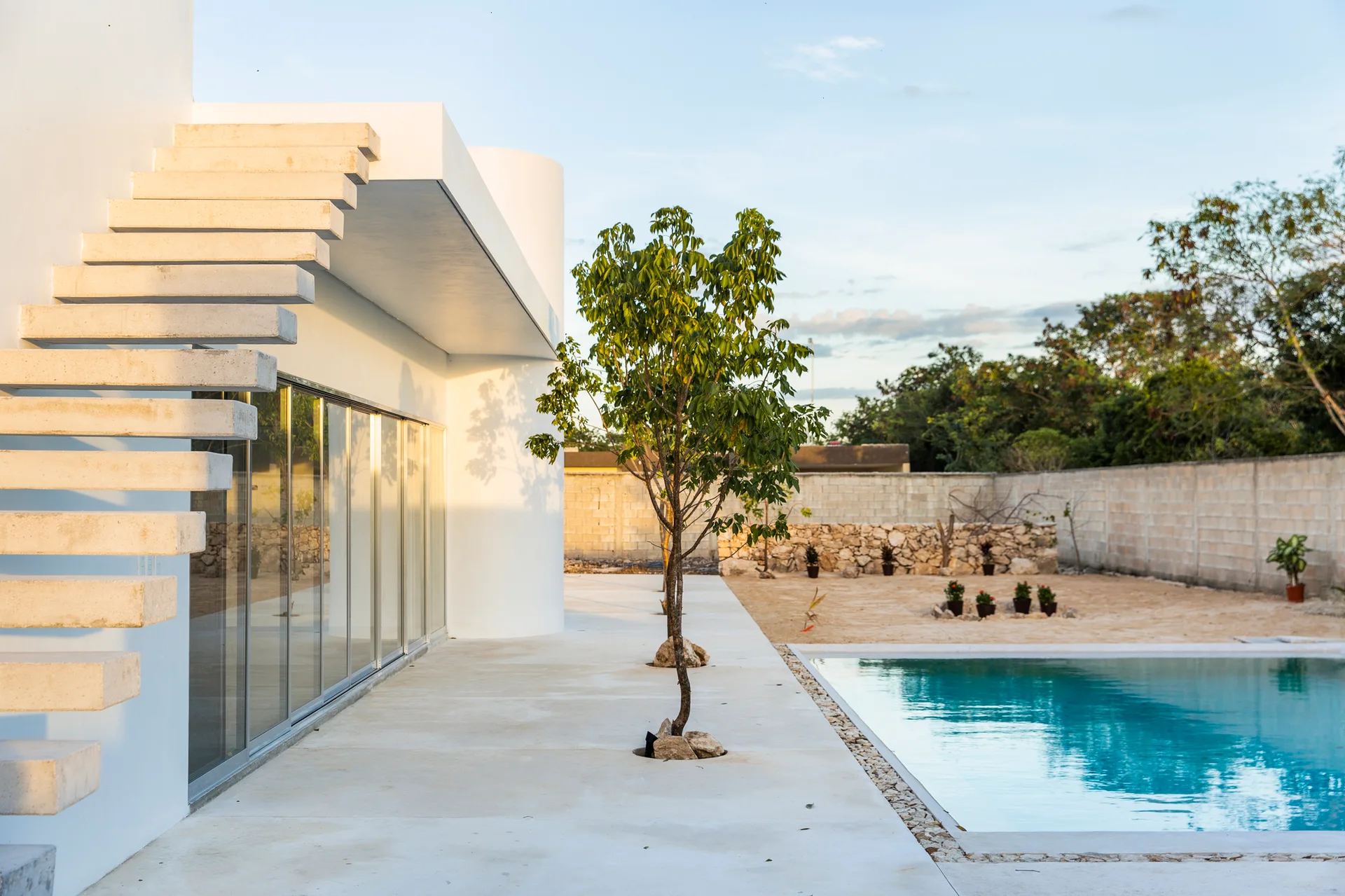A minimalist white stucco building features a cantilevered concrete staircase next to a concrete pool deck and a rectangular swimming pool.