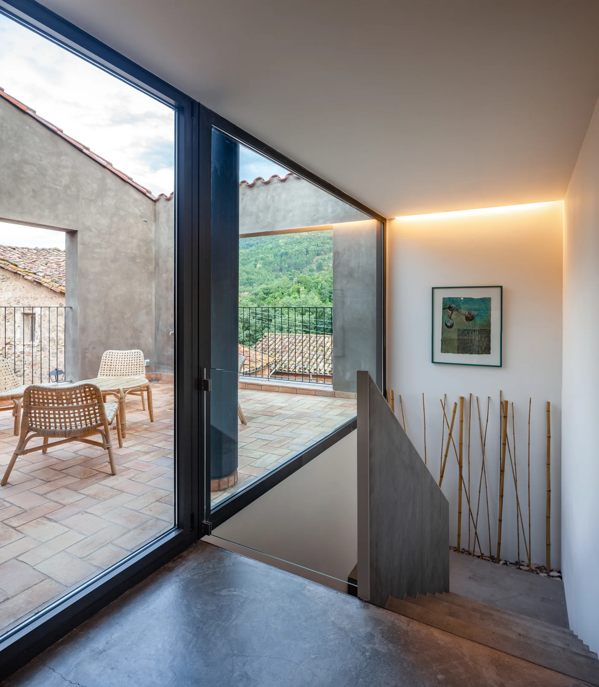 A concrete staircase landing features large glass sliding doors that open to an outdoor terracotta terrace with wicker chairs.