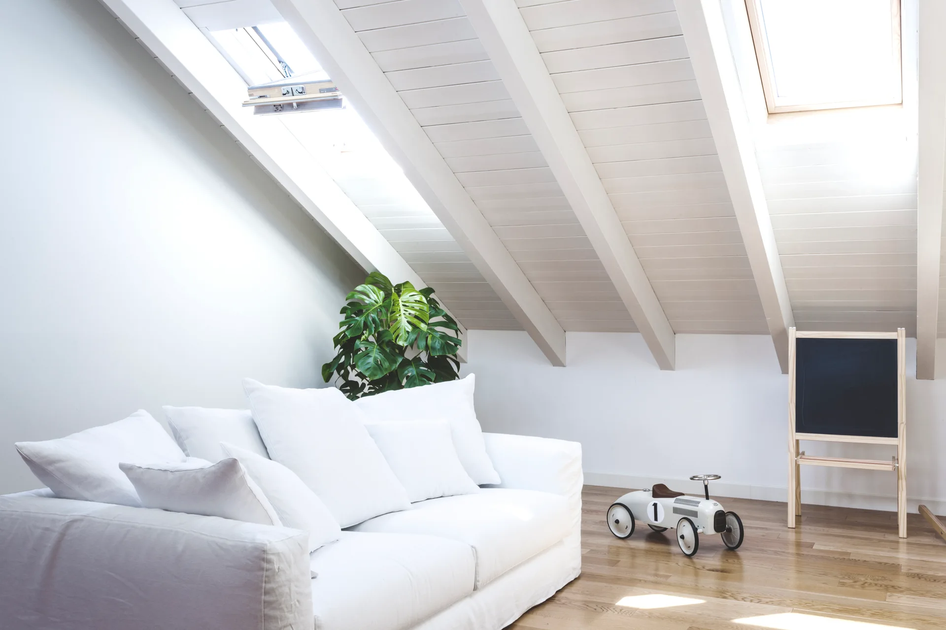 A bright attic living space featuring a white fabric sofa, skylights, painted wood ceiling beams, wood flooring, and a vintage toy car.