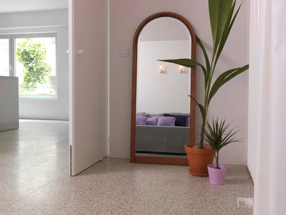 An entryway featuring a large arched wooden floor mirror next to potted plants on a speckled terrazzo floor.