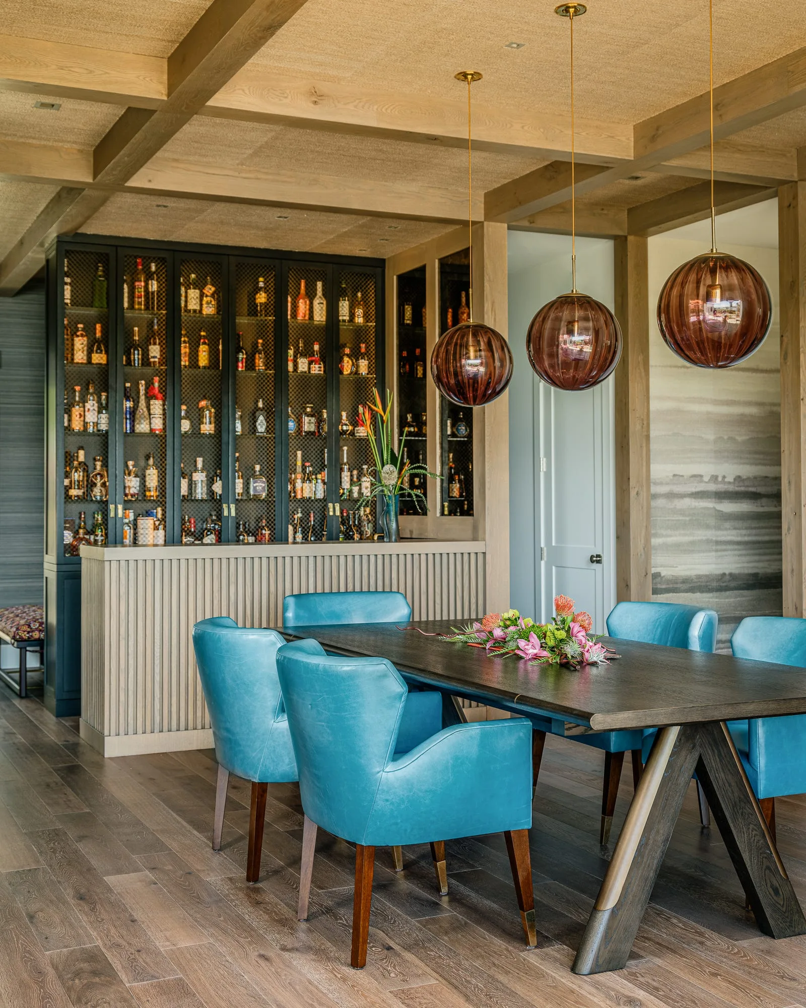 A dining room features a dark wood table surrounded by turquoise leather chairs beneath three large glass globe pendant lights and a coffered ceiling.
