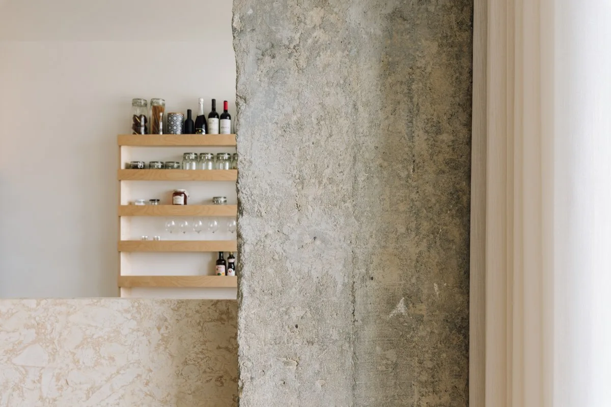 A textured concrete pillar stands beside a natural wood pantry shelving unit stocked with glass jars and bottles behind a stone partition.