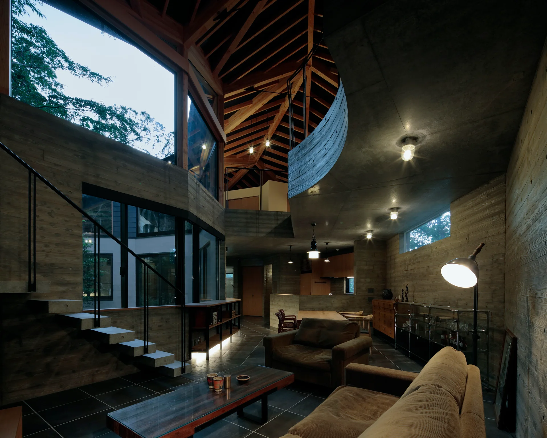 Modern living room featuring board-formed concrete walls, a floating staircase, two brown sofas, and an intricate exposed timber ceiling structure.