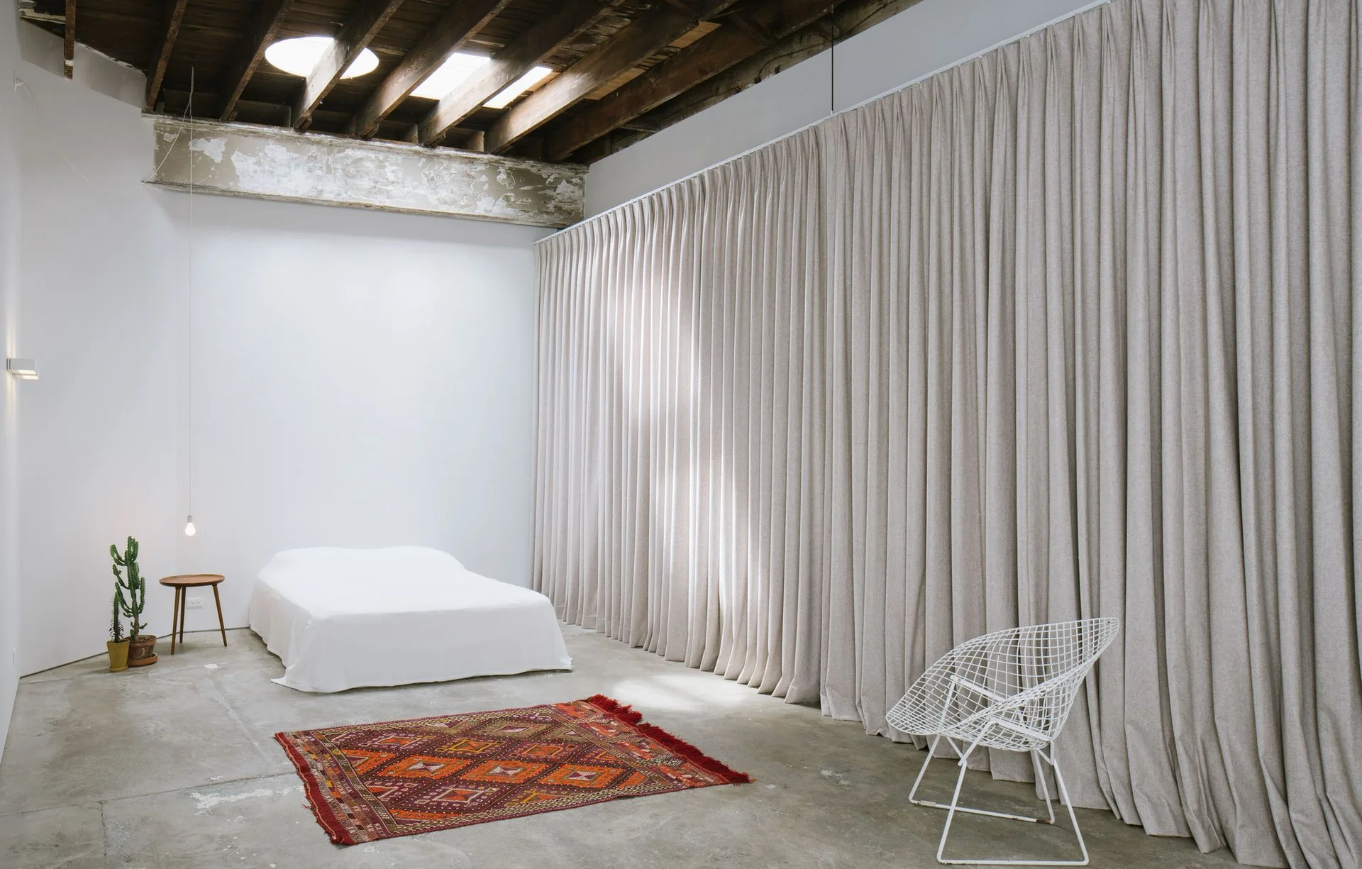 A minimalist loft bedroom features a low bed with white linens, a large beige curtain wall, exposed wooden ceiling rafters, and a concrete floor.