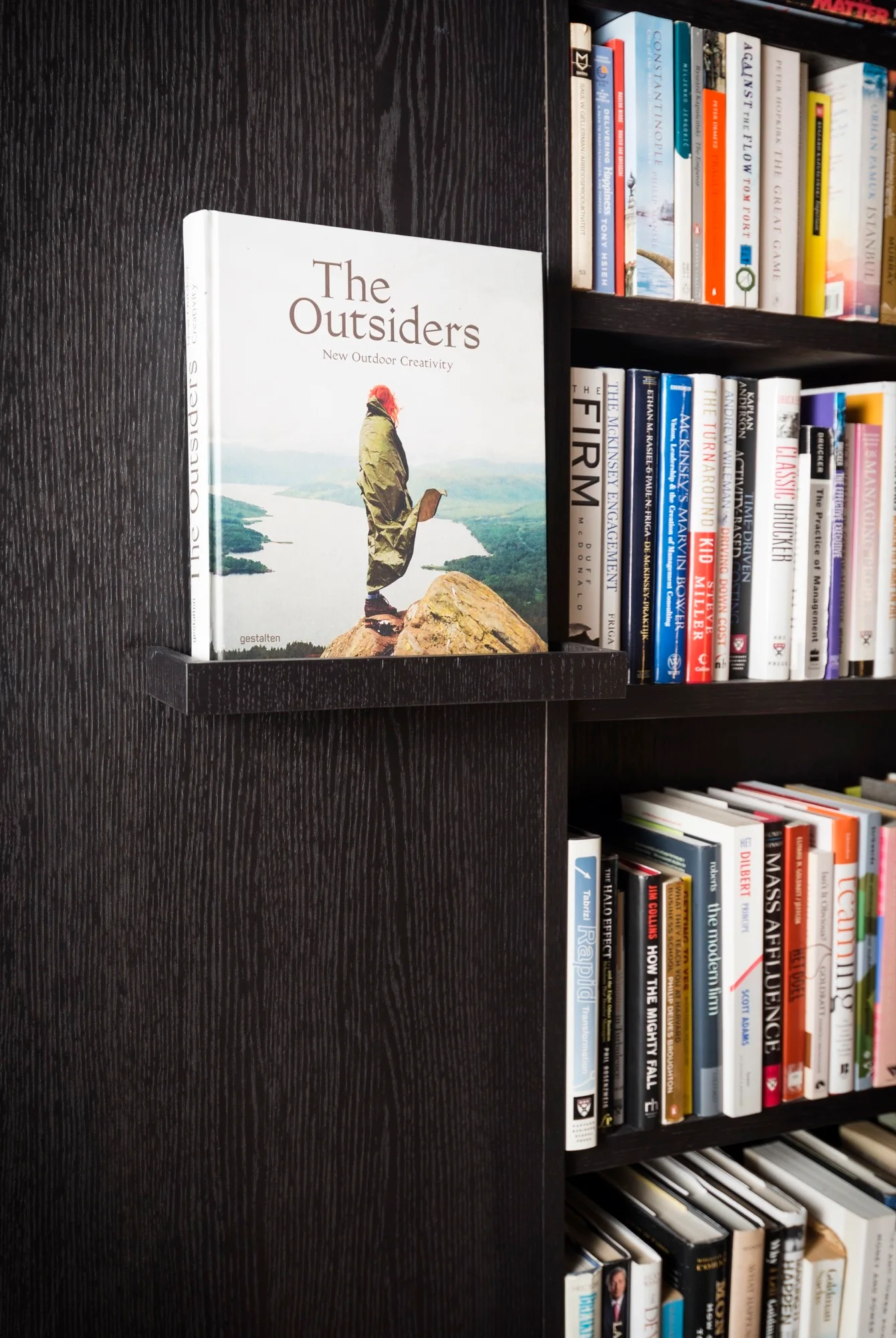 A dark wood grained bookshelf filled with colorful books, featuring a single book displayed cover-forward on a small integrated ledge.