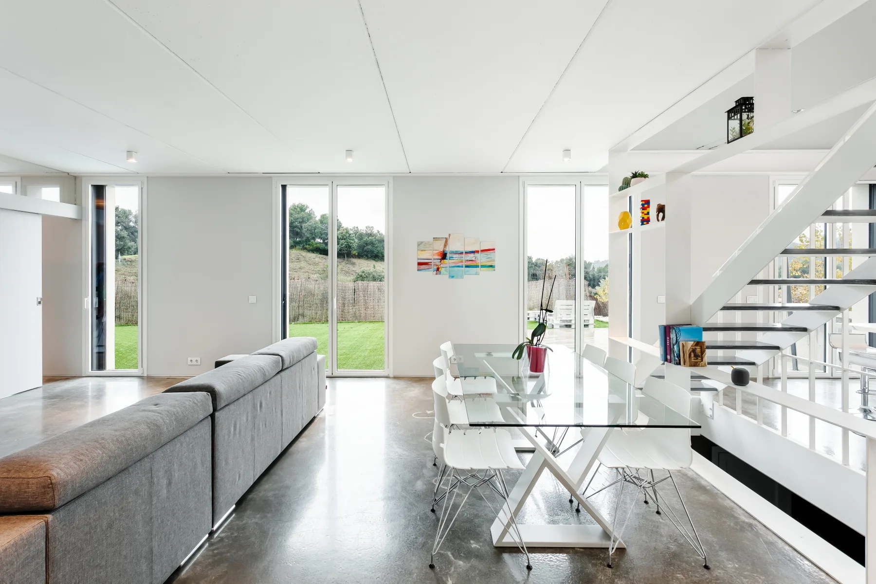 An open-plan living and dining area with a grey sectional sofa, a glass-topped dining table with white chairs, and a minimalist white staircase.