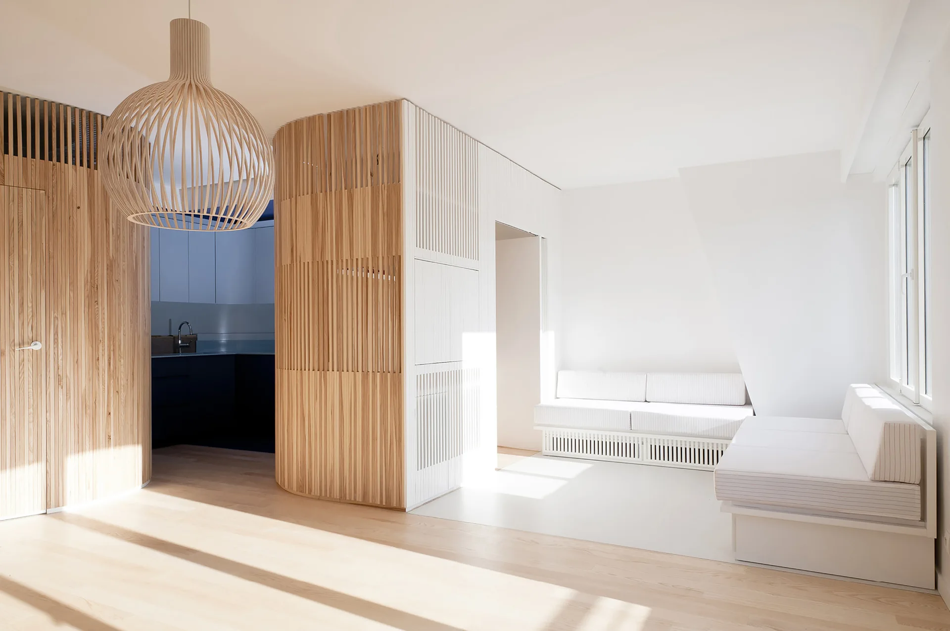 A minimalist bedroom featuring light wood plank flooring, a curved wood slat partition, white platform beds, and a matching slatted pendant light.